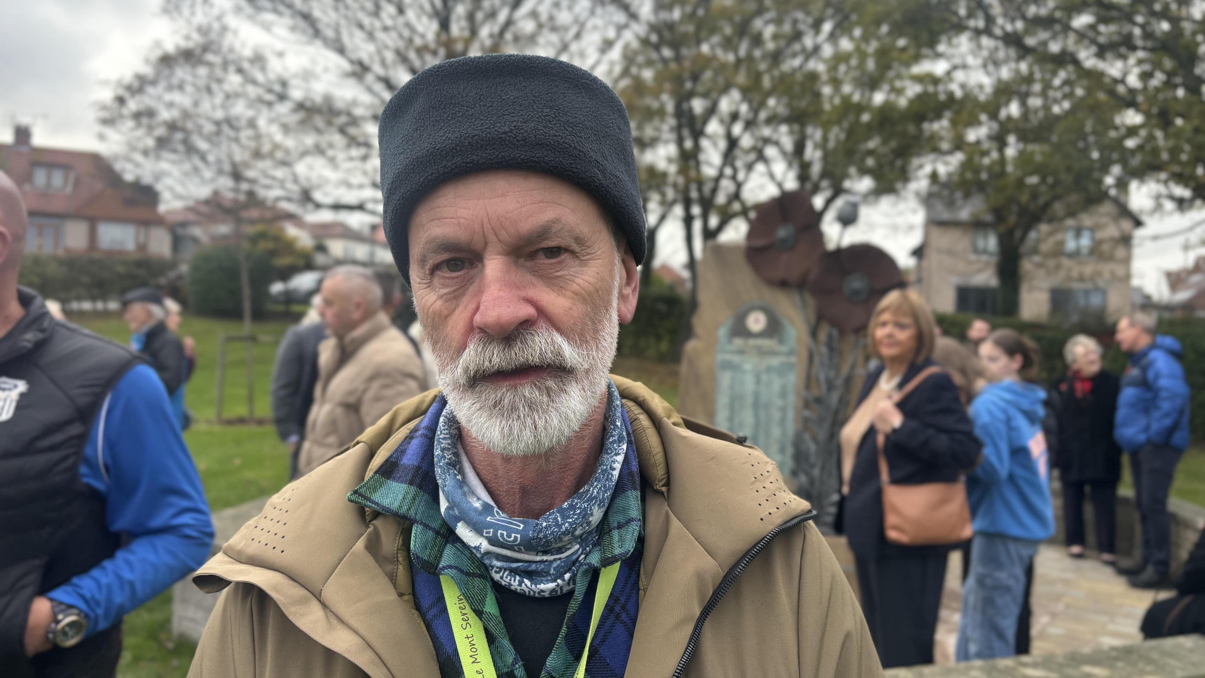 Colin Wilbourn has a white beard and a black hat. He is wearing a brown jacket. Behind him is the poppy sculpture and people standing in groups. They are standing in a park. Mr Wilbourn is not smiling at the camera.