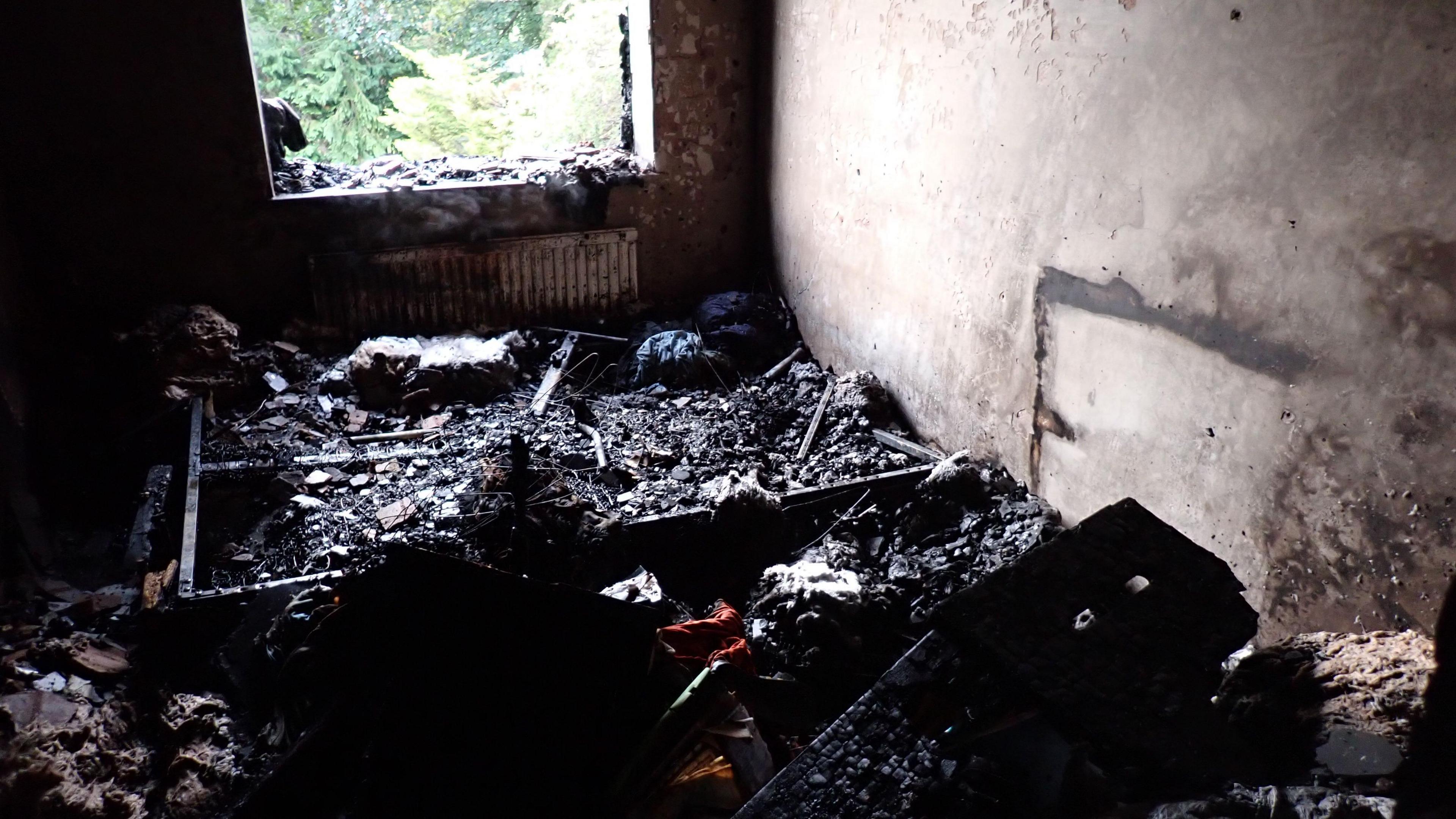 A room that has been totally burned down by a fire. There is a window on the wall and underneath a radiator, but everything else is burnt and black and the walls are smoke-stained.