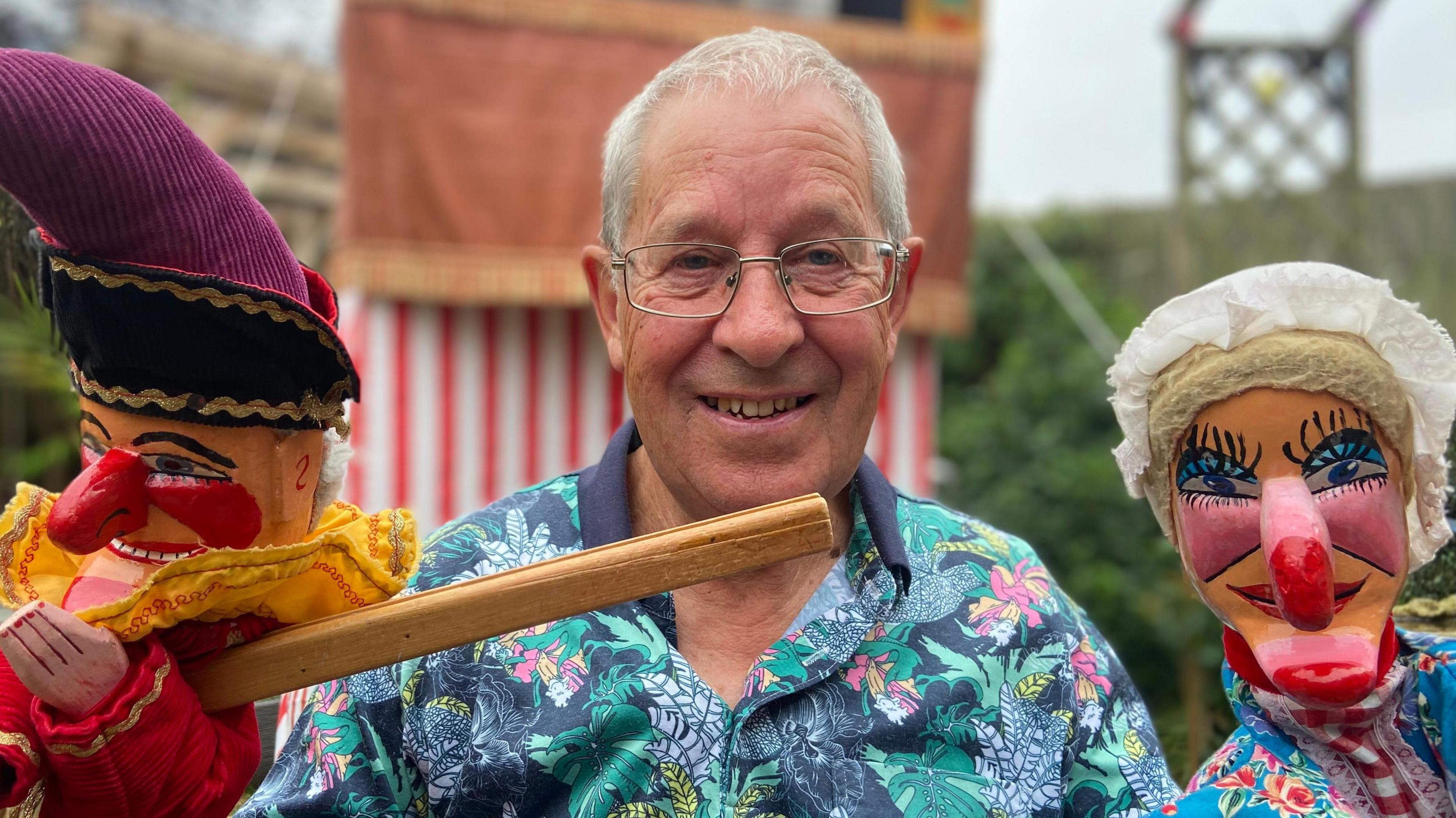 Paul Douglas. who has short grey hair and wears glasses and a bright floral shirt, smiles as he demonstrates his traditional Punch and Judy puppets, with his red-and-white stall in the background. 