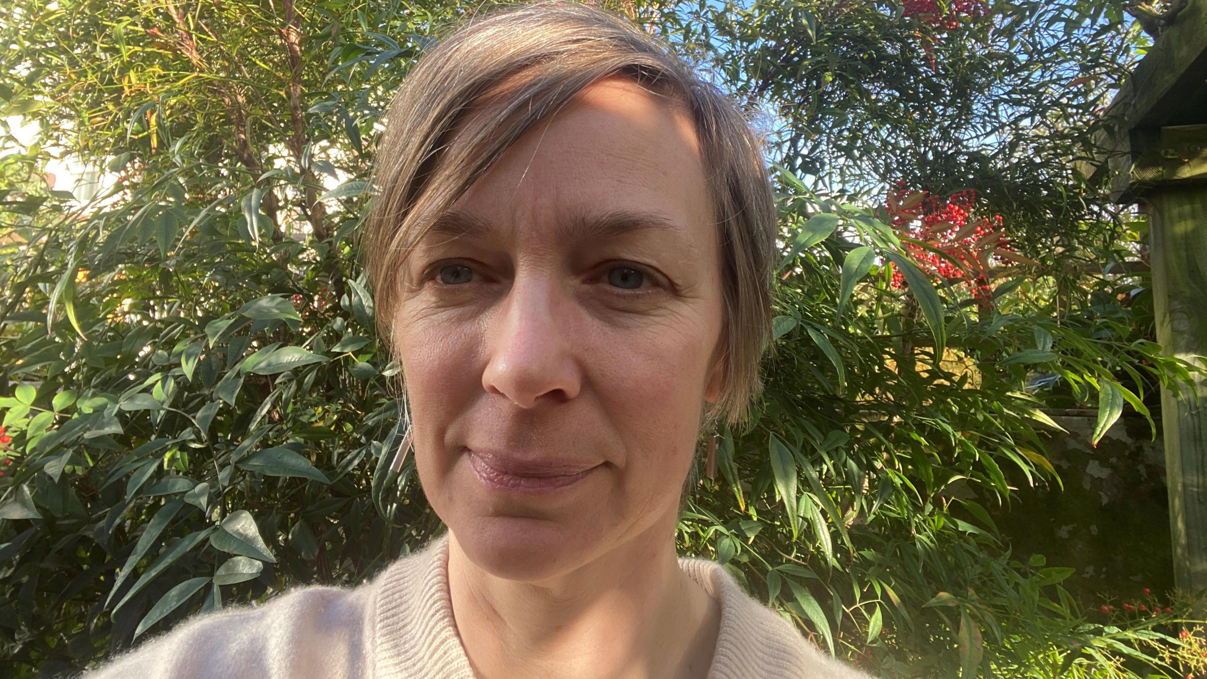 Selfie of Fiona Button in a garden, head and shoulders only. She is 48, has short blonde hair and is wearing a beige wool jumper.