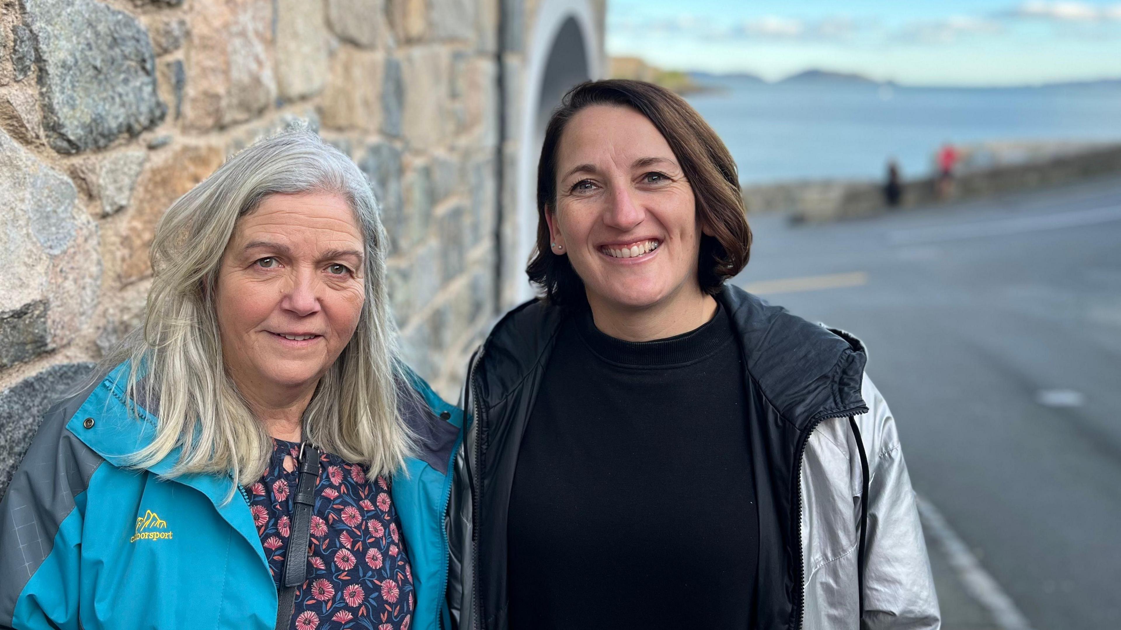 Anne Wainwright and Liz Stonebridge stood on the pavement at the bottom of the Val Des Terres. Liz is wearing a black top and silver jacket. Anne is wearing a blue jacket and floral top. 