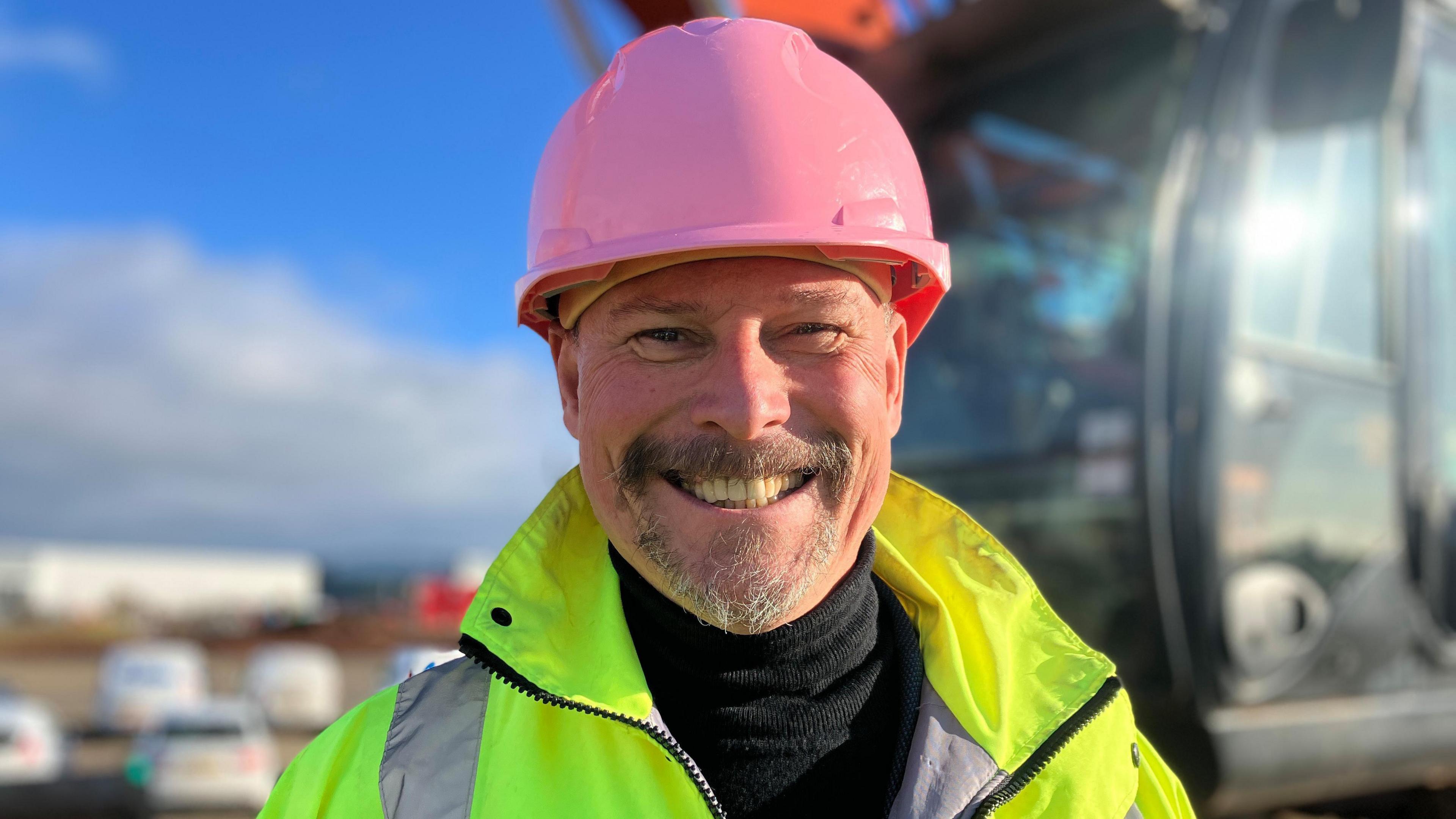 A man with a goatee-style beard wearing a black rollneck jumper, a hi-vis jacket and a pink helmet smiles at the camera.