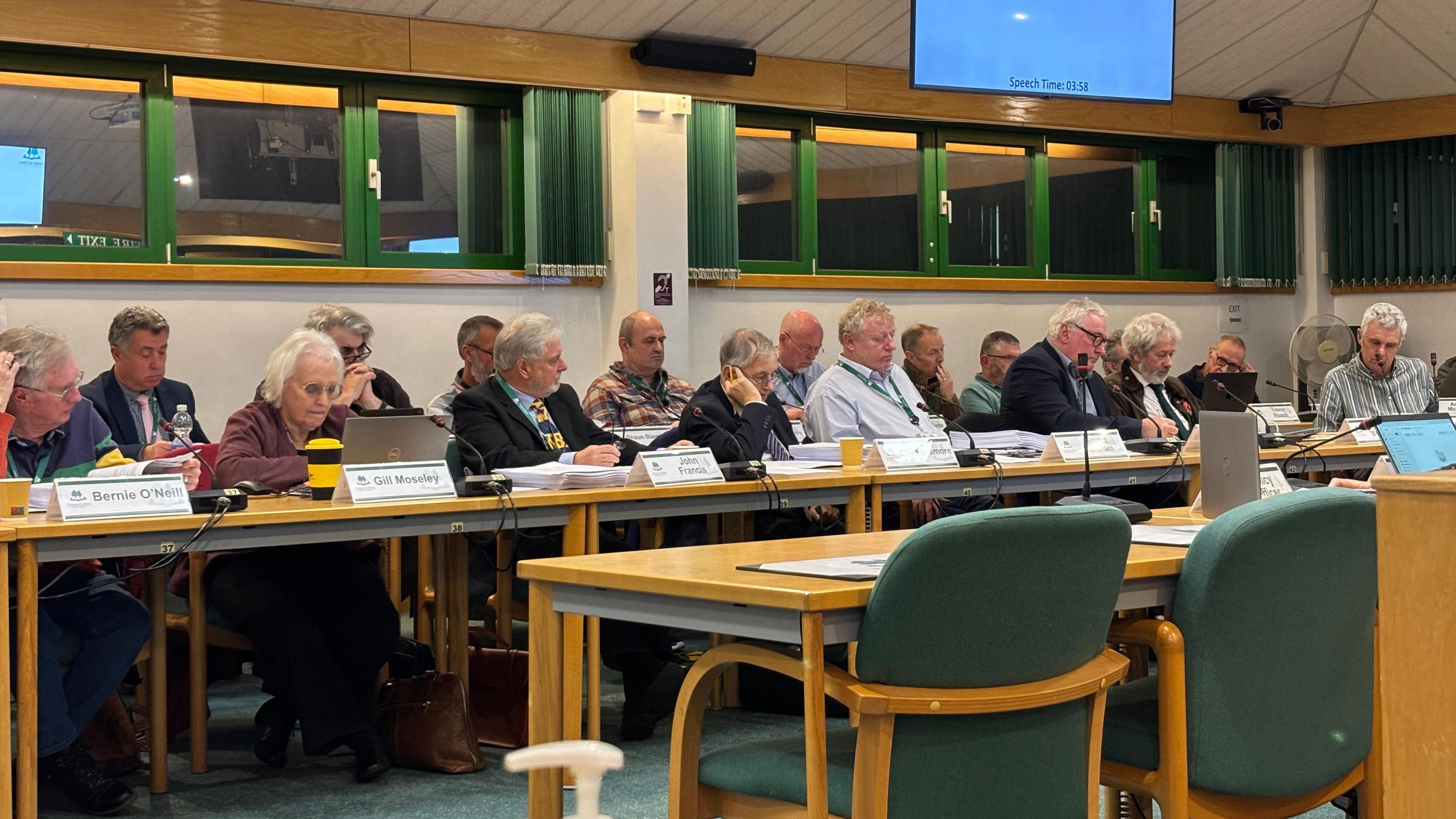Two rows of councillors sit in a meeting at Forest of Dean District Council with name tags in front of them In the foreground are two green chairs against the desk facing the councillors.