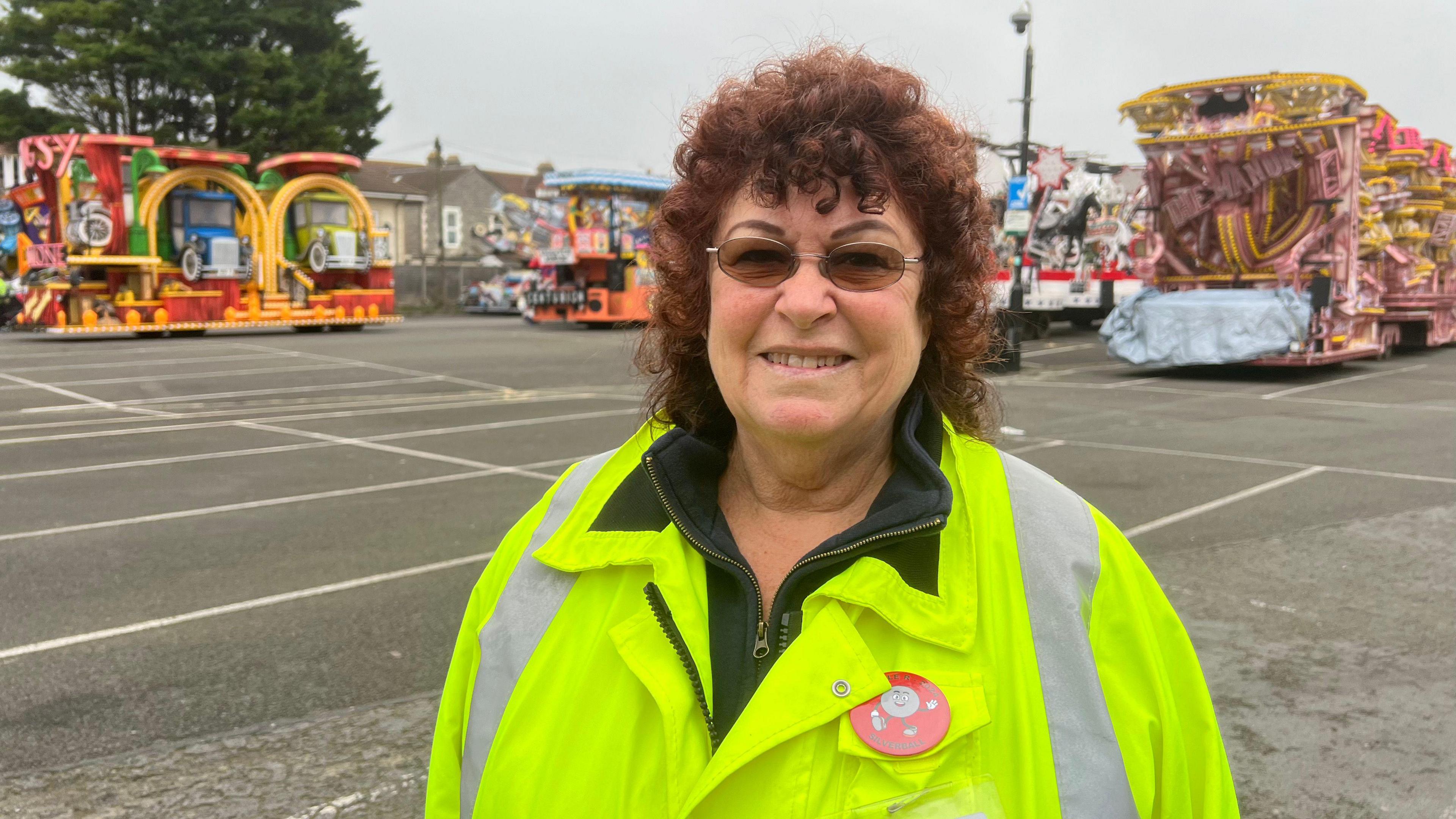 Head and shoulders image of Jan Matthews who has curly brown hair and is wearing glasses and a hi-vis jacket. She is standing in front of parked carnival carts.