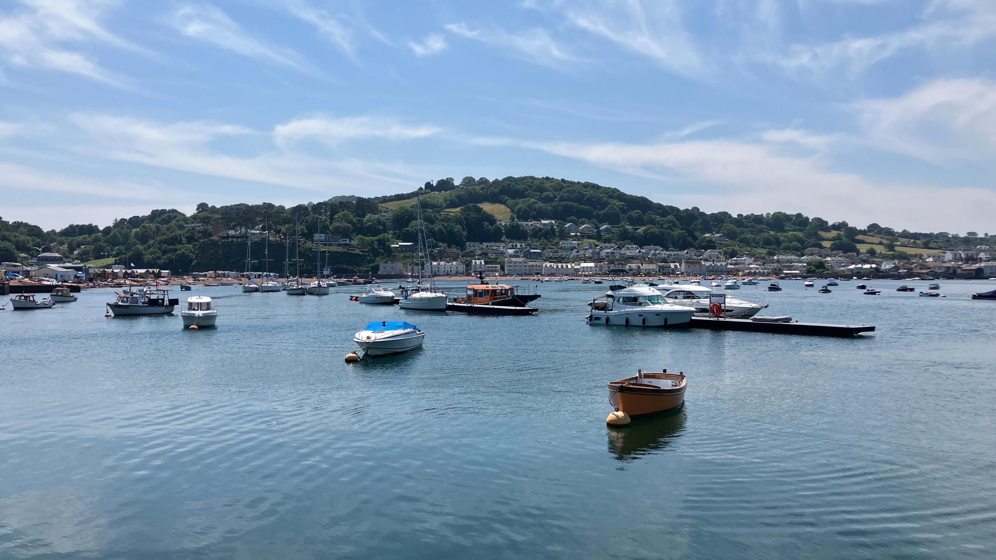 A wide shpt of a river estuary on a sunny day. There are small and medium sized boats moored in the water. there are wisps of cloud in the sky. the far shore of the river is visible, with buildings along the edge of the water.