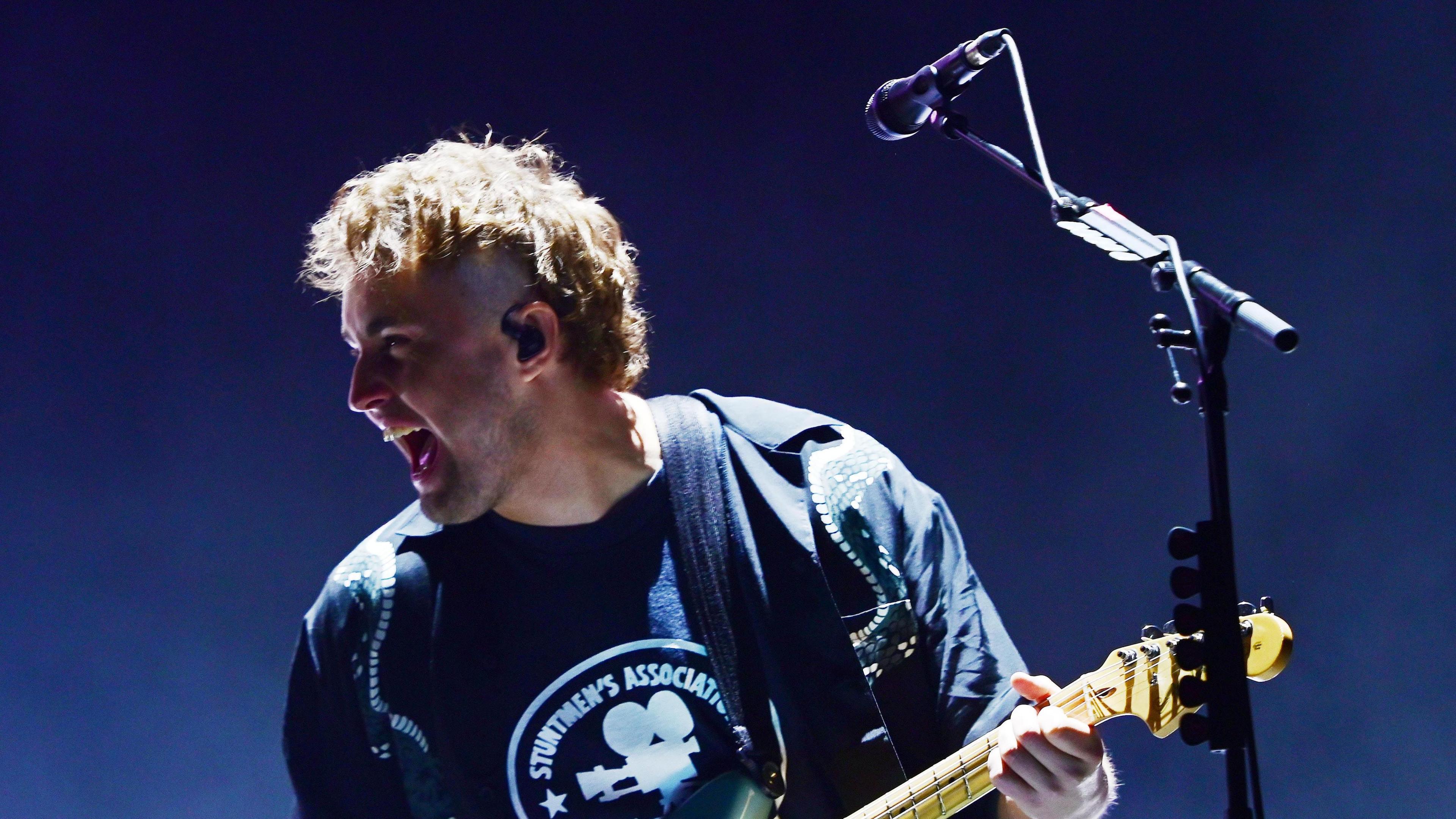 Sam Fender playing guitar next to a microphone on a stand. He is grinning and wearing a navy t-shirt.