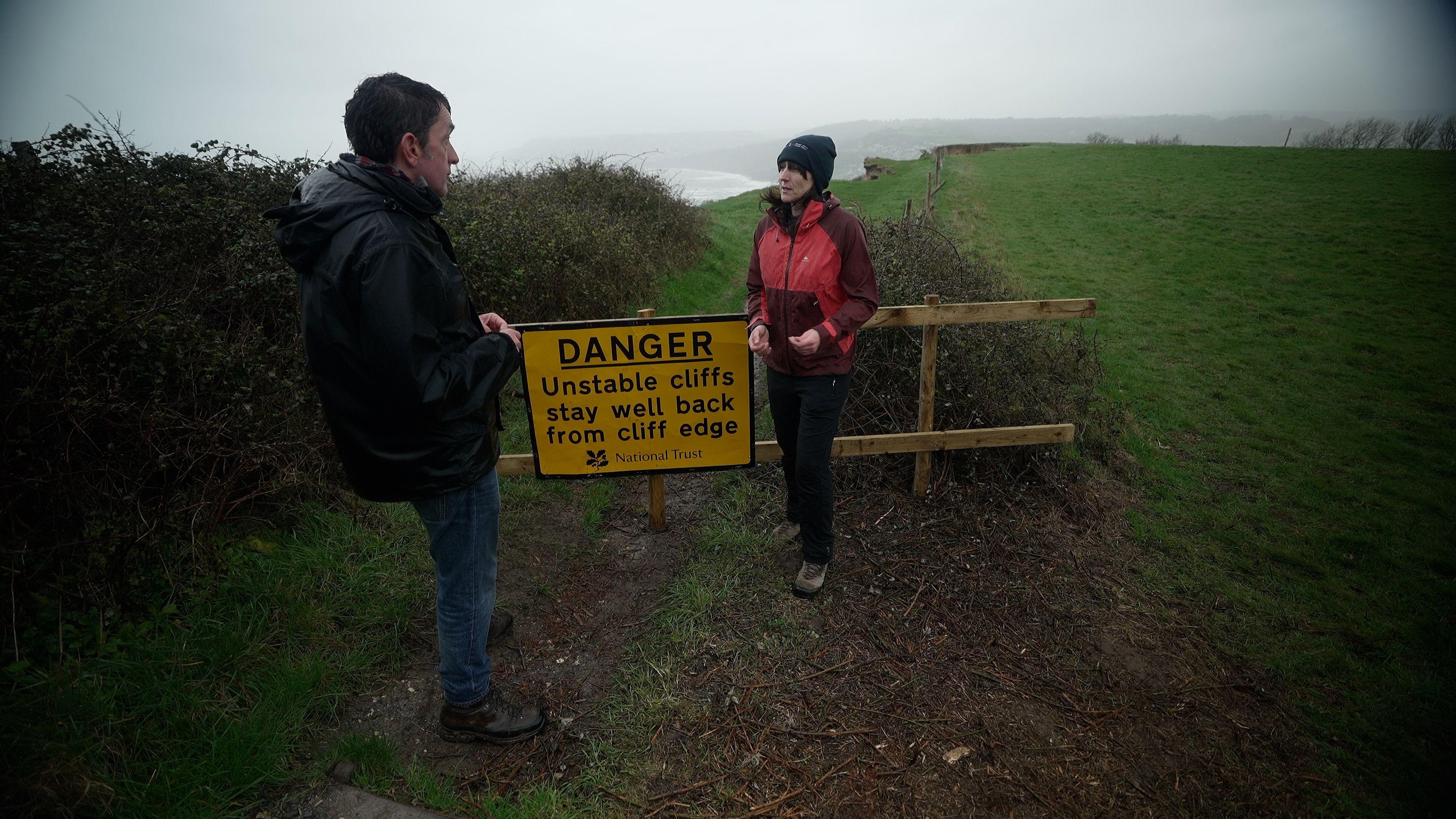 Lorna Sherriff from South West Coast Path Association stands with the reporter, Justin Rowlatt, by a National Trust yellow sign saying danger - unstable cliffs, stay well back from cliff edge. Both look west and the sky and the sea in the background are grey. The path is closed with a wooden gate just behind where they are standing. A cliff edge is seen a few meters after the wooden gate.