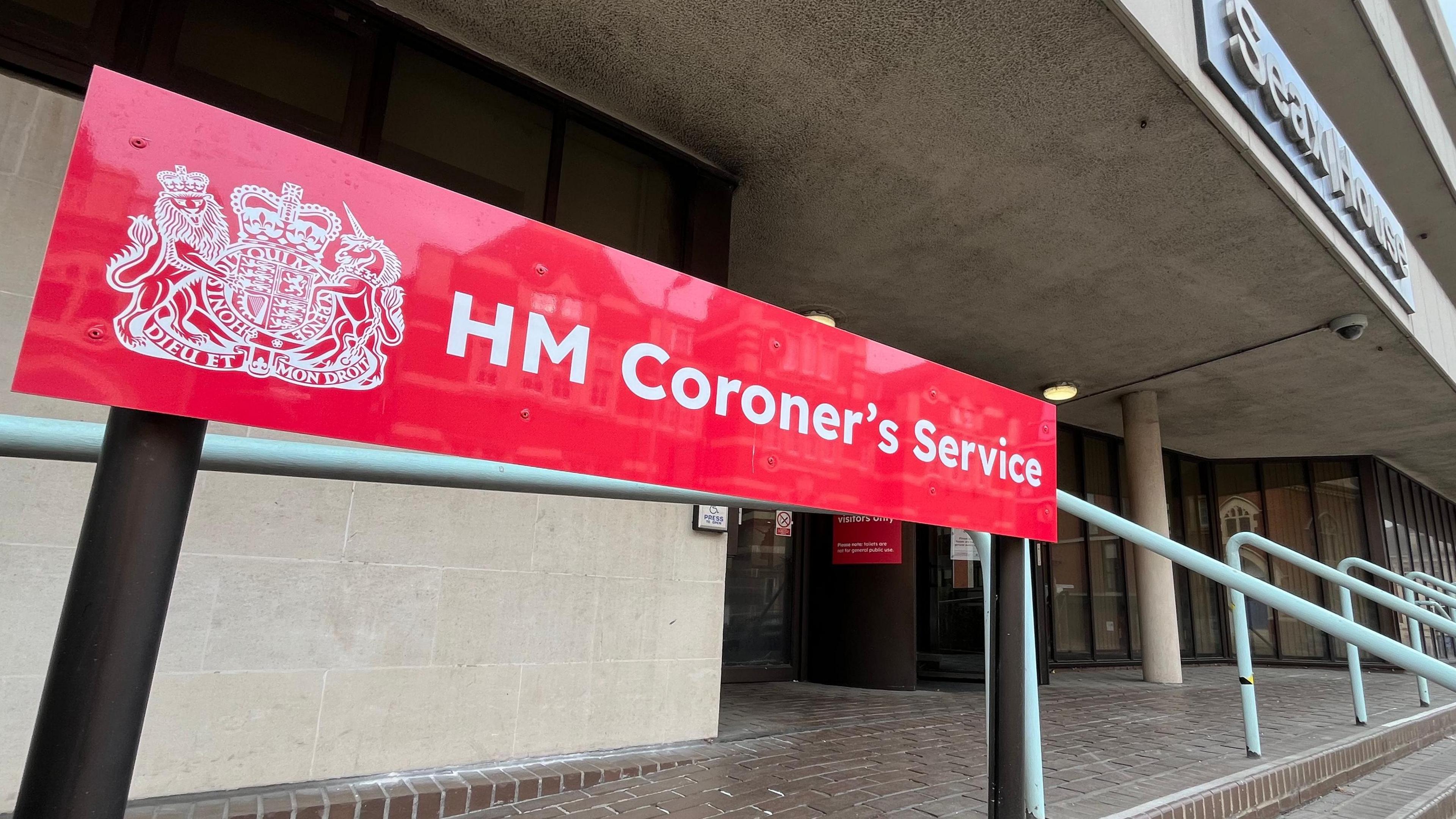 A red sign at the entrance to the coroner's court, which says "HM Coroner's Service". There is a brick path outside the entrance, which is a revolving door, and steps lead to this point.