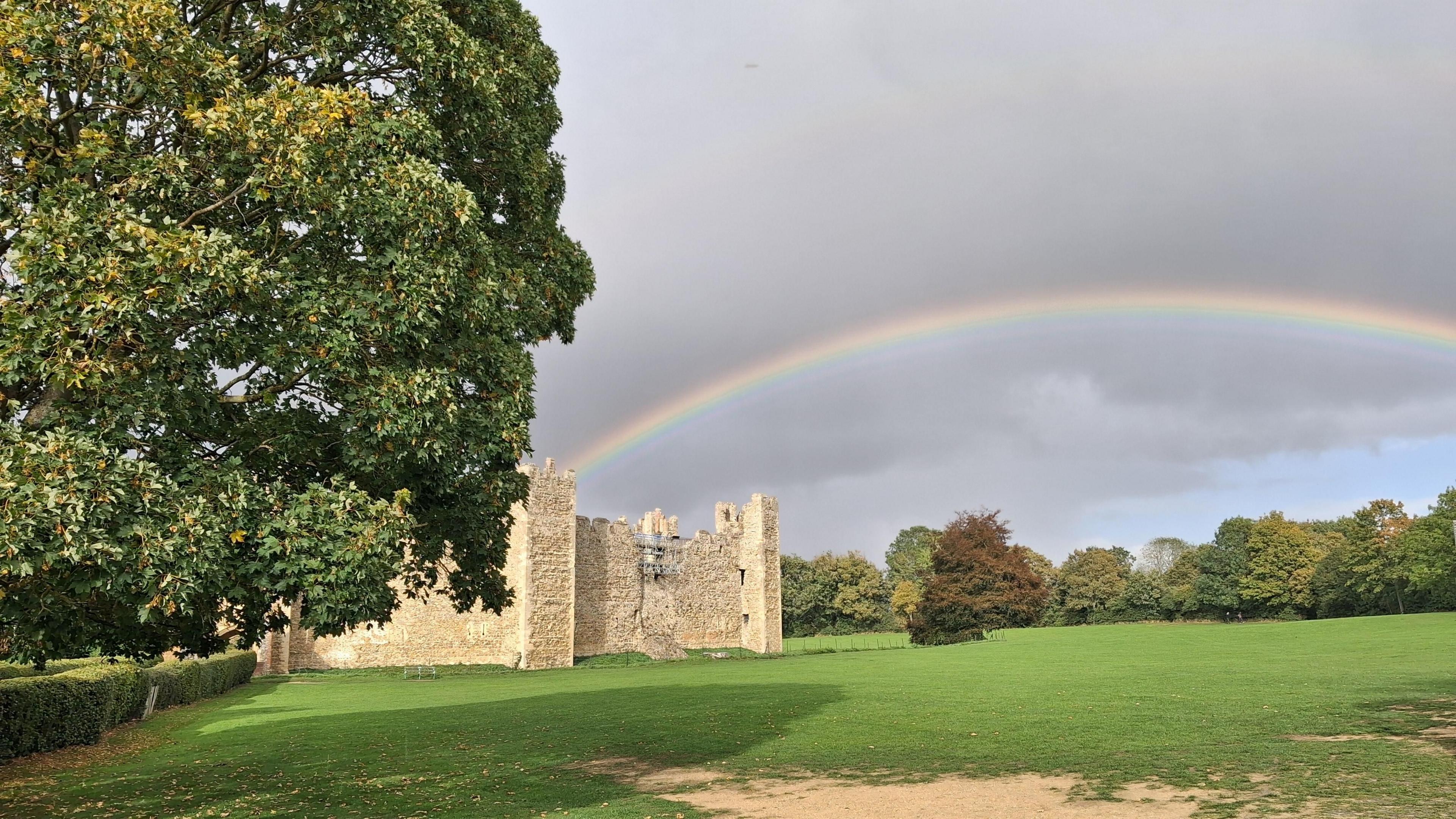 A castle in a grass field. A large rainbow can be seen above it in a grey cloudy sky. A large tree can be seen to the left of the image. 