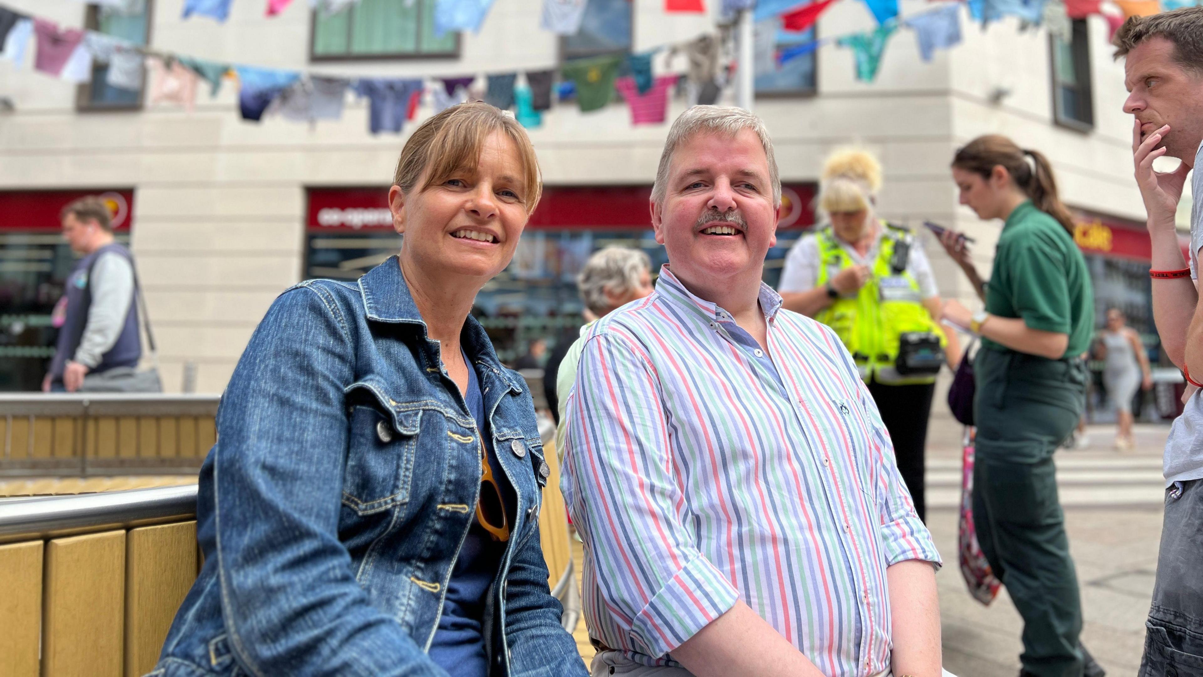 Alix and Andrew sit on a bench and smile at the camera with the shirts display in the background