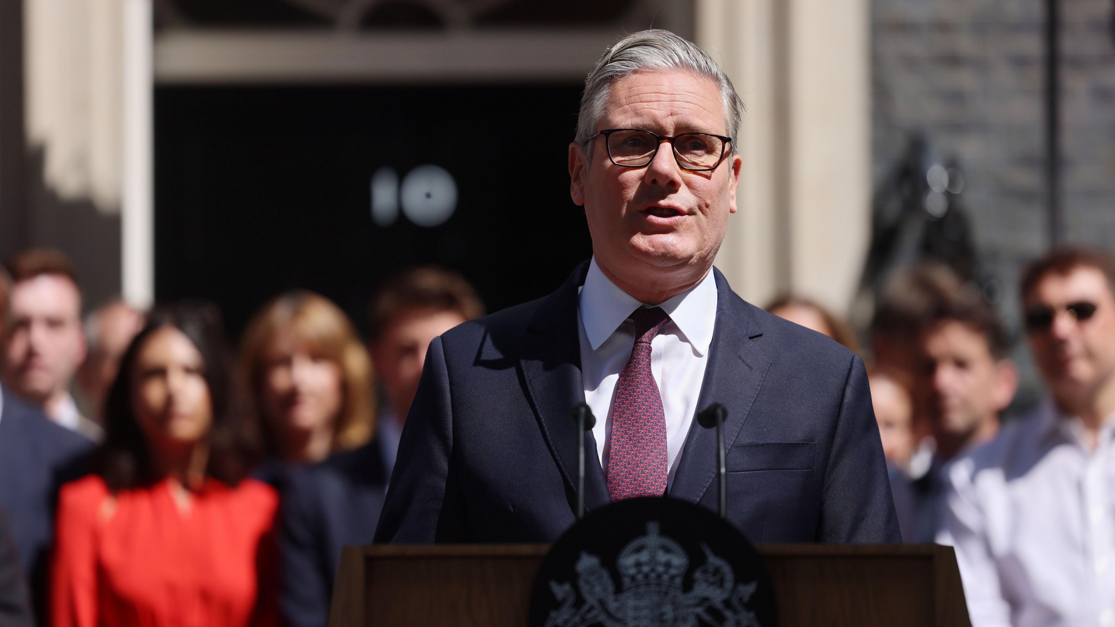 Sir Keir Starmer stands outside of Downing Street delivering a speech. He wears a black suit, red tie and glasses.