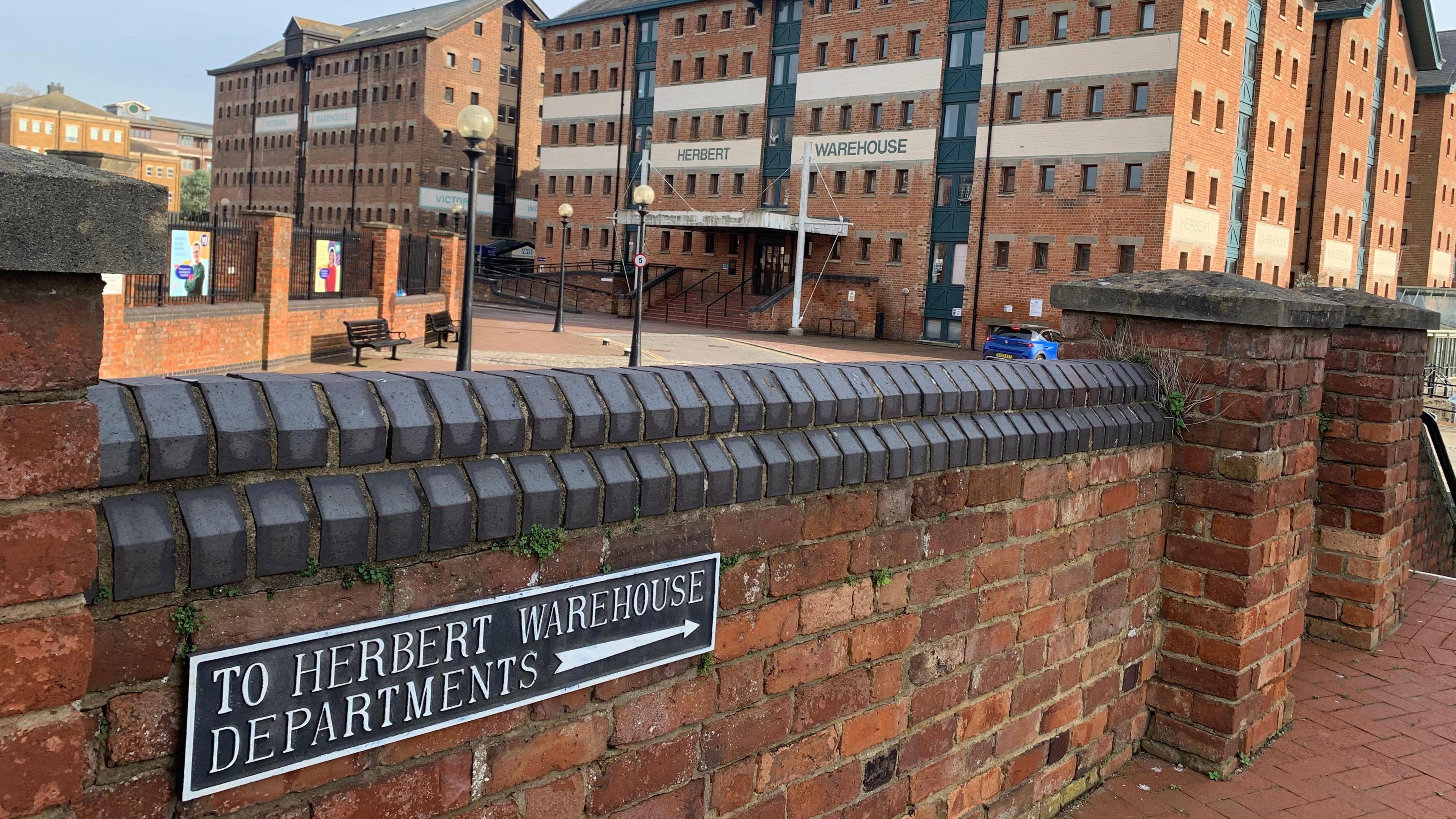 Herbert, Kimberley and Philpott warehouses at Gloucester Docks. It is a red brick building with a short brick wall in the foreground.