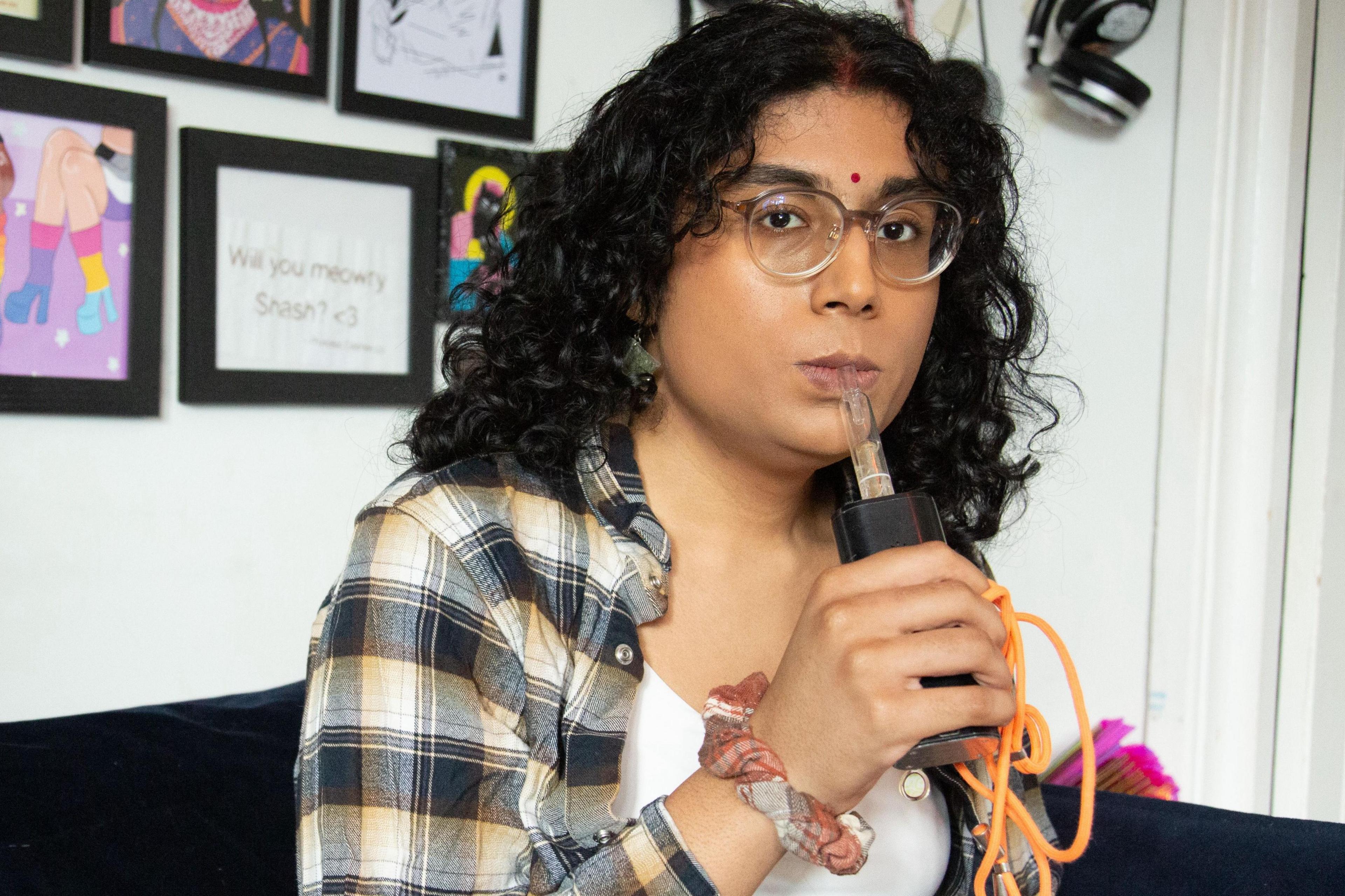 A woman with curly long dark hair and clear rimmed glasses puffs on a large cannabis vaporiser with an orange lead trailing out of it