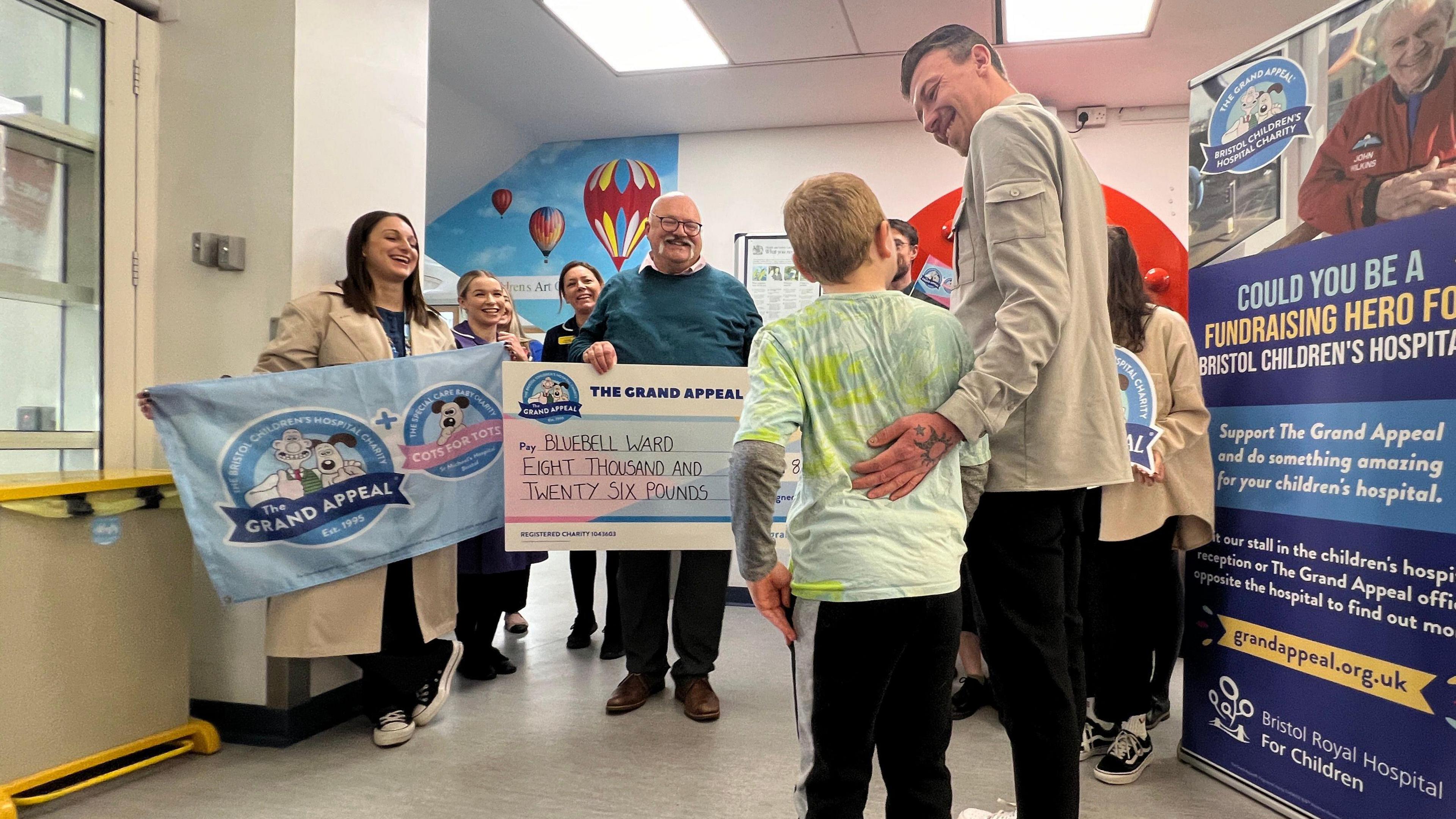 Harry (left) and father (Edward) seen smiling at each other as Bluebell Ward staff hold a giant cheque and smile at them. 