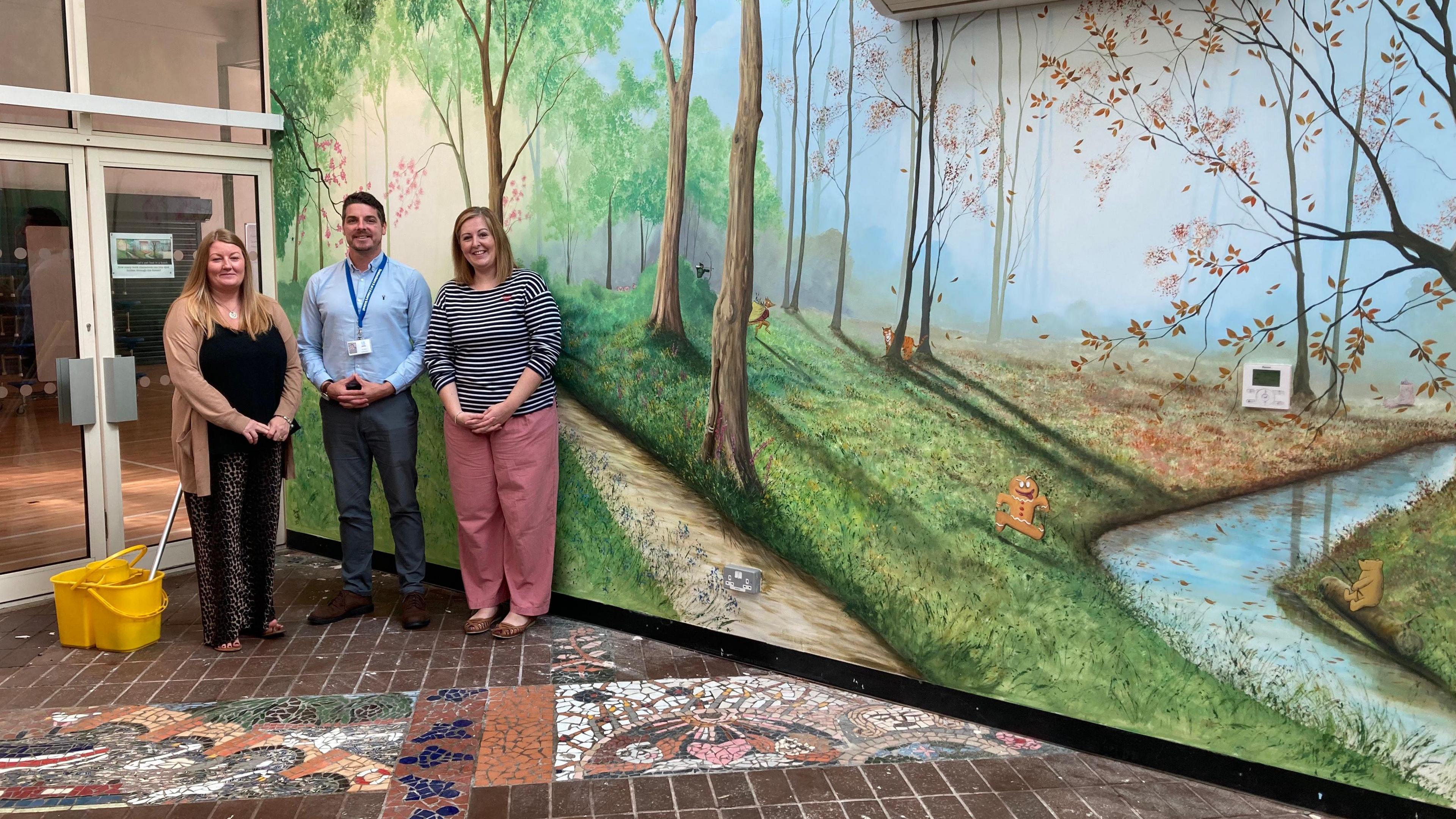 Philippa Moore, Tim Rylatt and Amanda Hebden standing in front of a mural at Tewkesbury C of E Primary School. They are standing in a line, smiling at the camera. There is a yellow mop bucket on the floor next to them. 