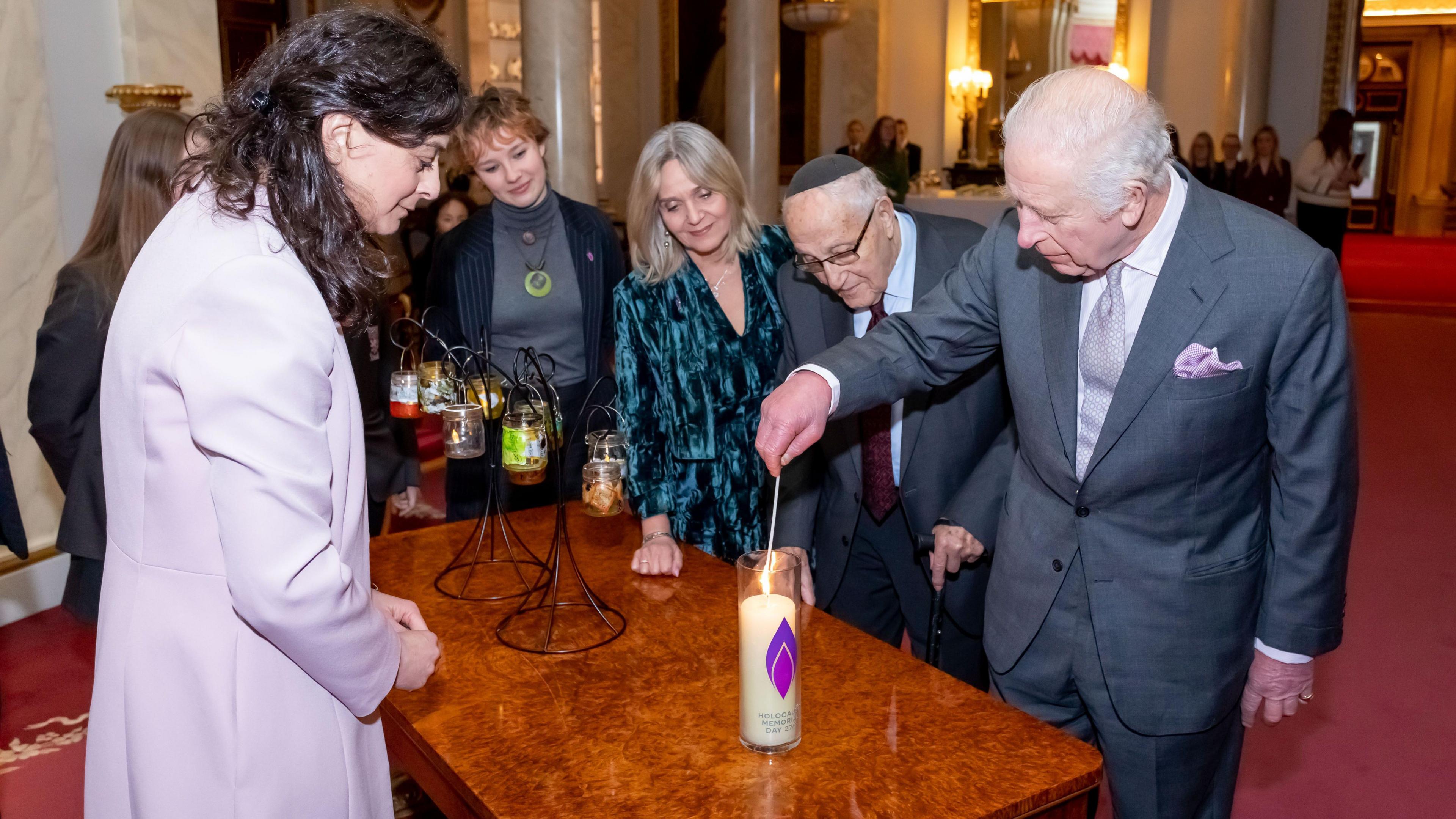 King Charles, wearing a grey suit, lights a candle saying "Holocaust Memorial Day" on the side of it while a small group of other people watches on.