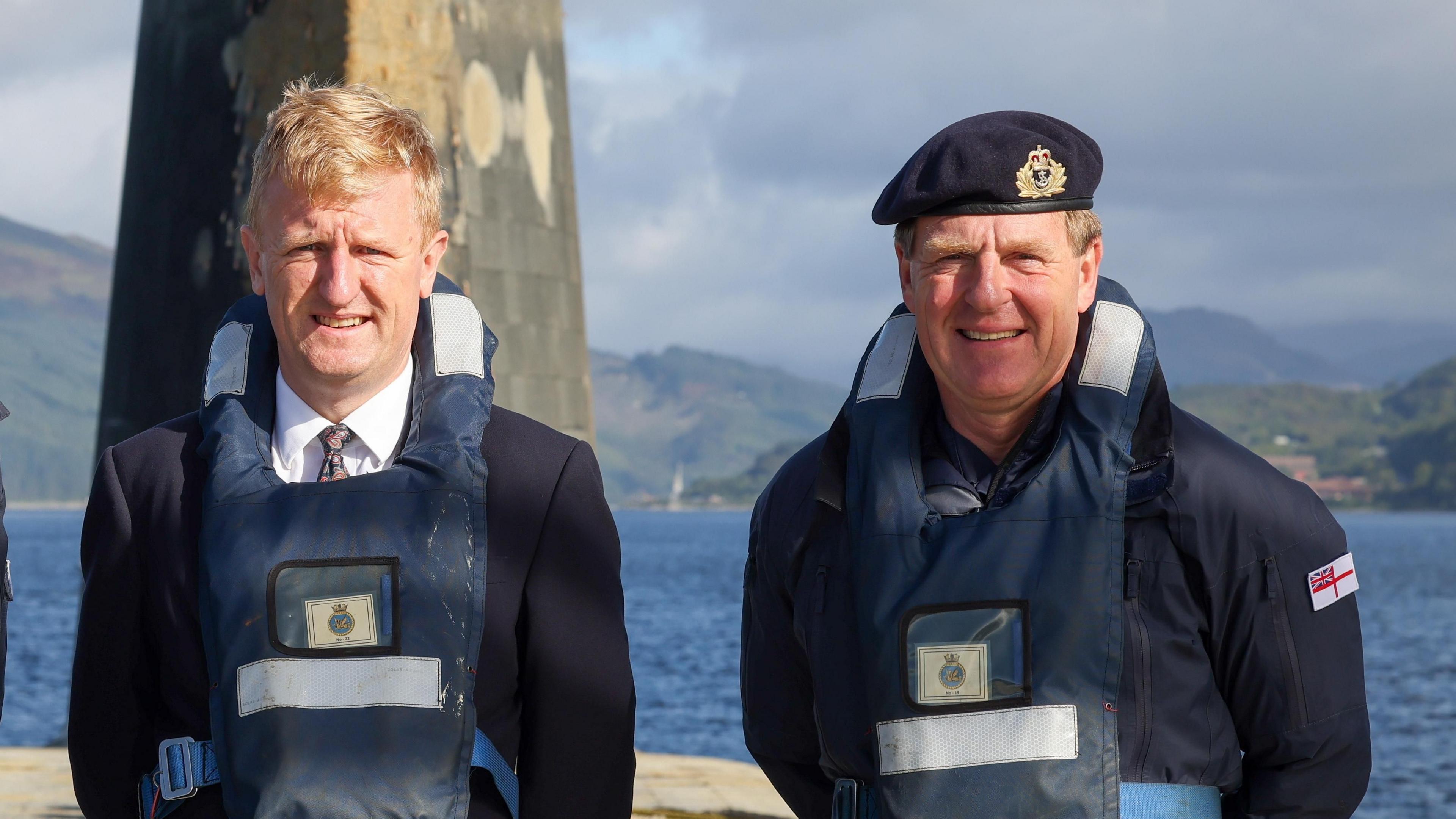 Oliver Dowden with Admiral Sir Ben Key, standing on a British Navy vanguard submarine with a lifeguard. Sir Ben is in uniform and a navy cap, Dowden is in a dark suit with a white shirt and patterned tie. 