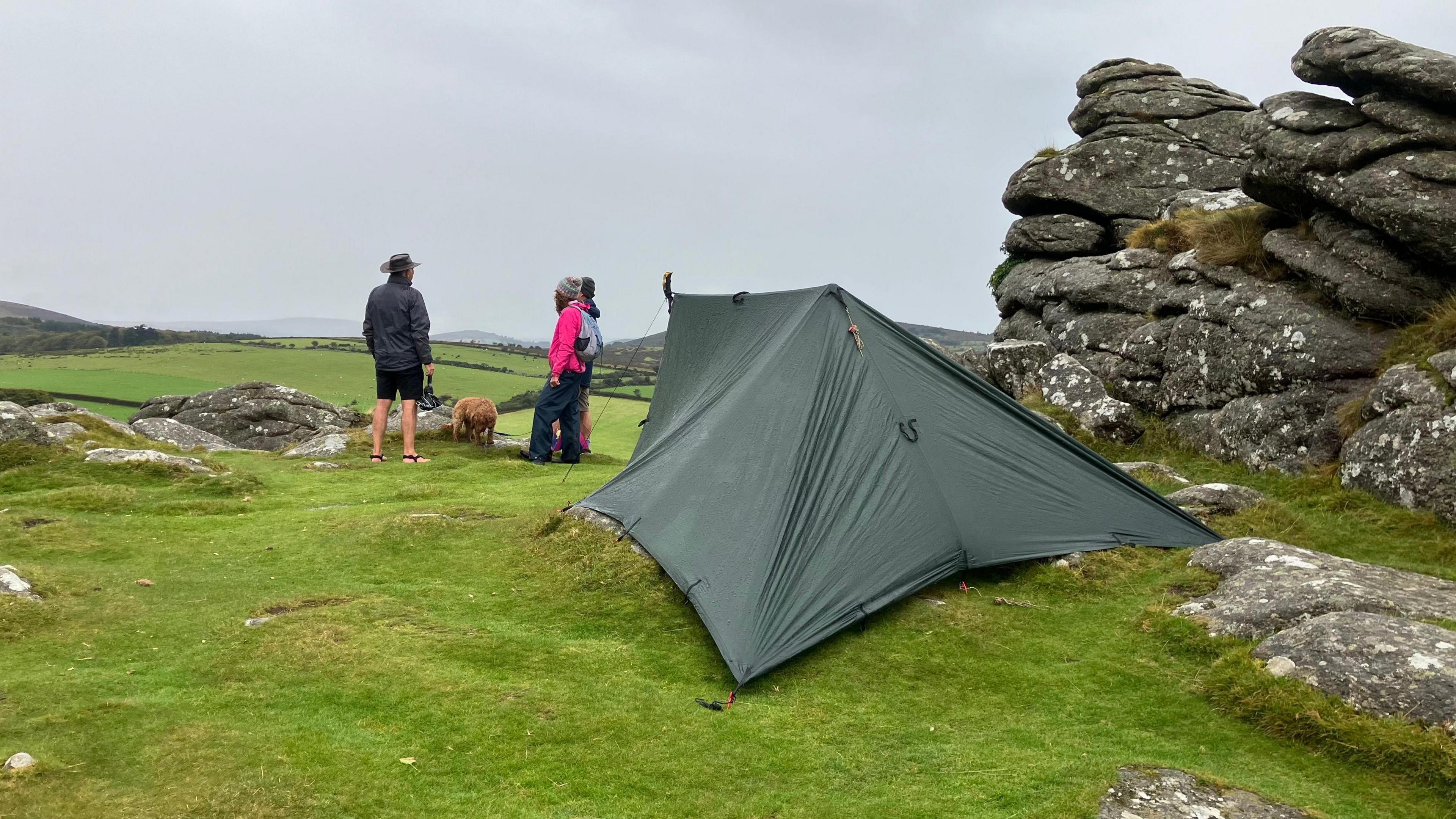 A green tent pitched next to a rock formation on Dartmoor. Behind the tent three people are stood talking to each other. One person has a brown dog on a lead.