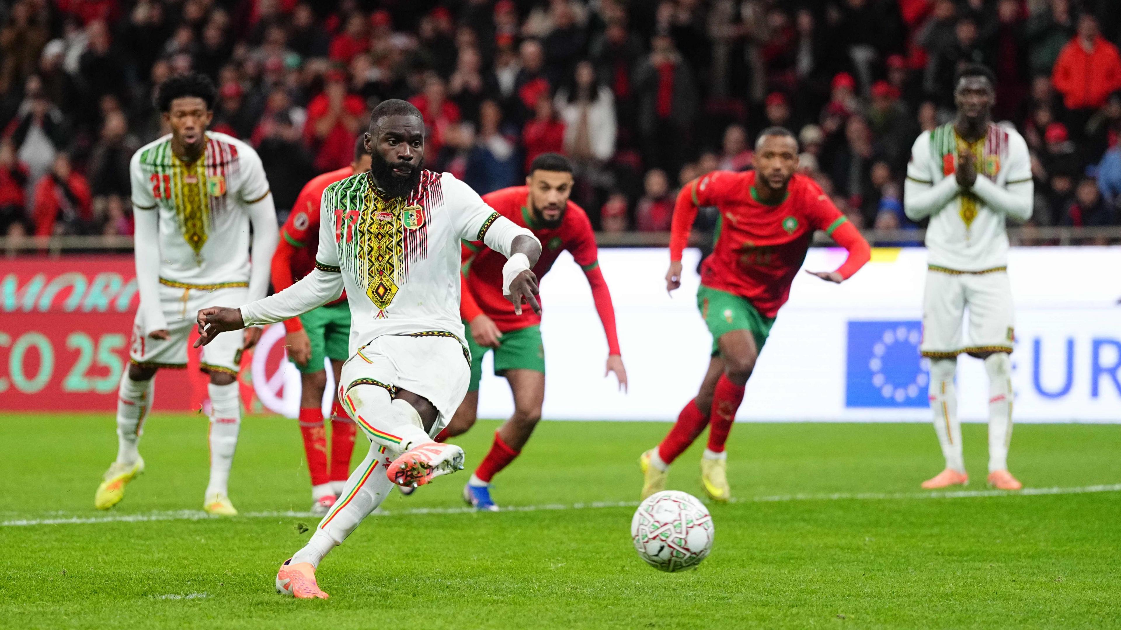 Lassine Sinayoko, wearing an all-white Mali kit, is seen mid-action as he scores a penalty at the Africa Cup of Nations against Morocco. Sinayoko has just kicked the ball, which is flying along mid-picture, just slightly elevated above the lush green turf. Five players are in the background on the edge of the penalty area, including two Mali players, one of whom has his hands clasped in a prayer gesture. Two of the three Moroccan players in red and green are already bursting into the box in anticipation