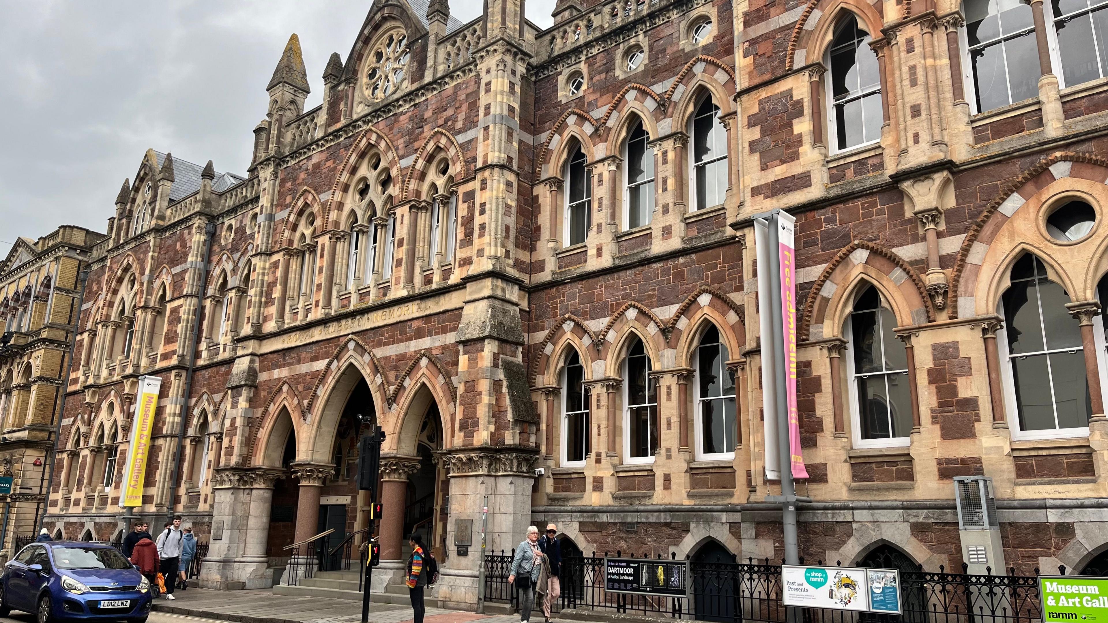 The exterior of the Royal Albert Memorial Museum in Exeter, which is in the style of Victorian Gothic Revival. The museum's exterior uses sandstone, has detailed stonework and uses characteristic Victorian features, including arched windows and ornate gables. People are walking past