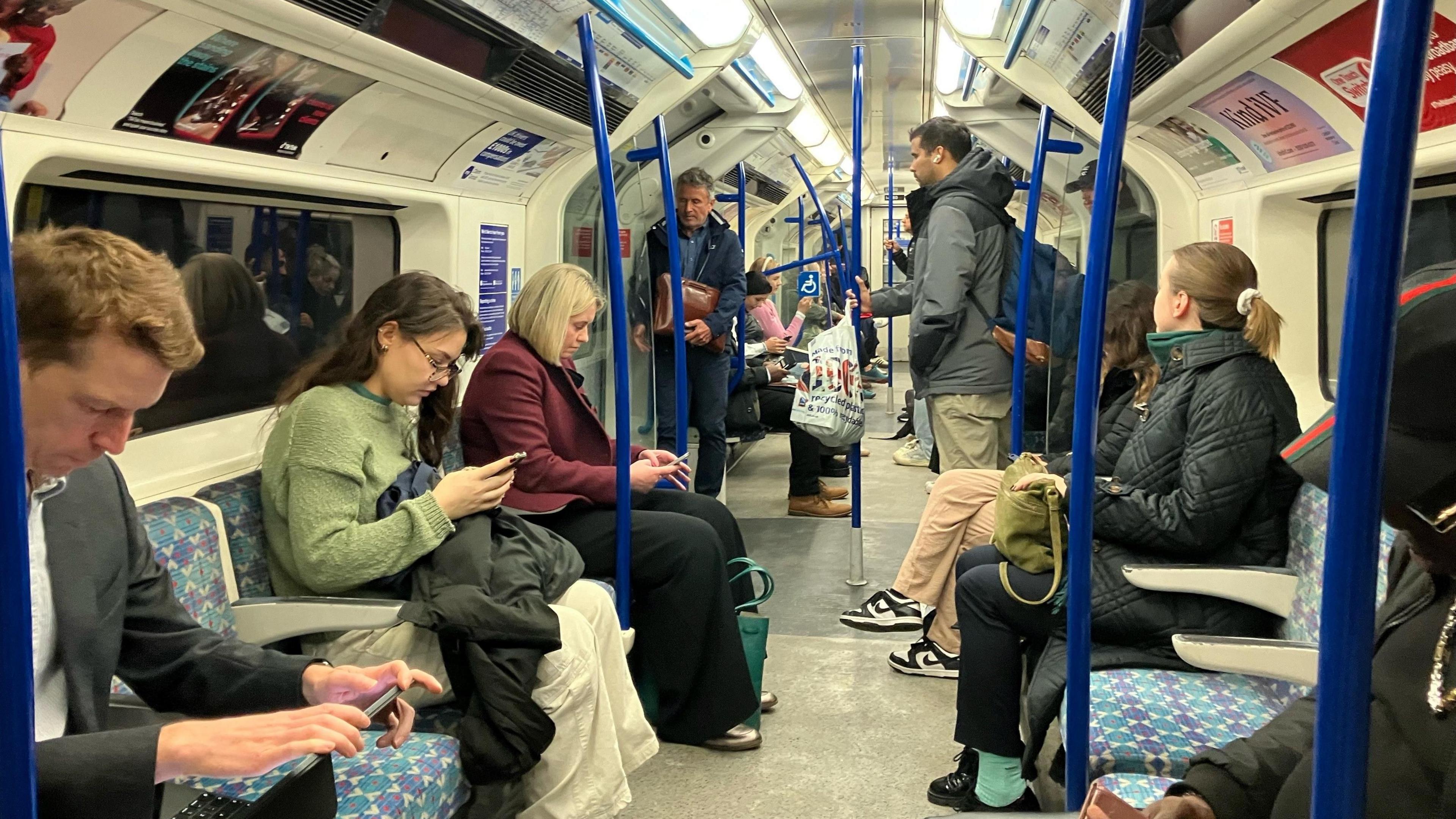 Passengers looking at their phones sitting down inside a Victoria line tube train, there are a few passengers standing holding the poles.