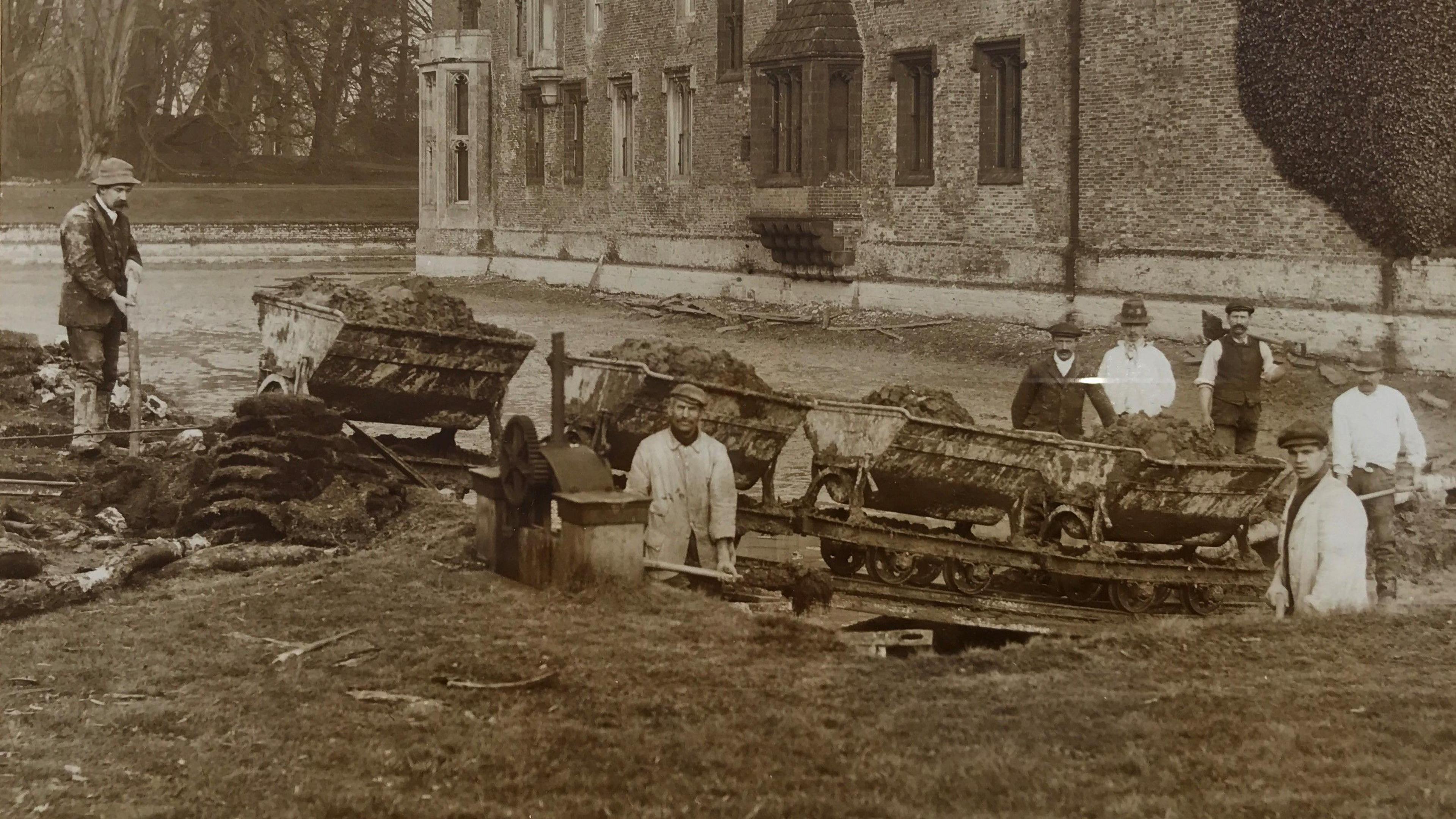 A sepia-coloured photo from 1910 featuring seven workers standing in and around the moat at Oxburgh Hall. They are wearing hats and some have spades. There are carts on wheels full of excavated earth.