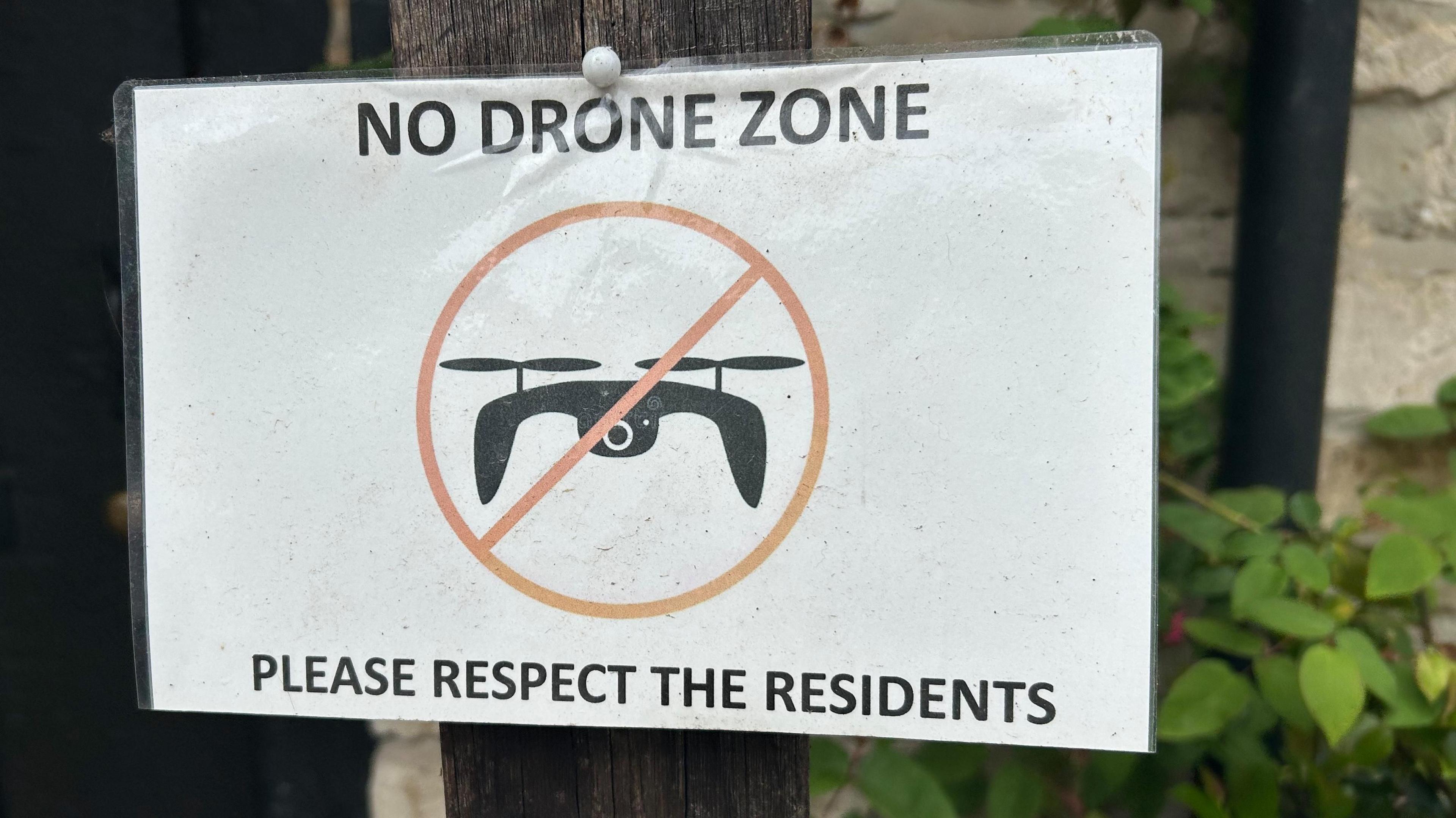 A white laminated piece of paper reads 'no drone zone, please respect the residents'. In the middle of the paper is a black drone graphic with a red cross in a circle over the top. The sign has been pinned to a wooden post outside a house.