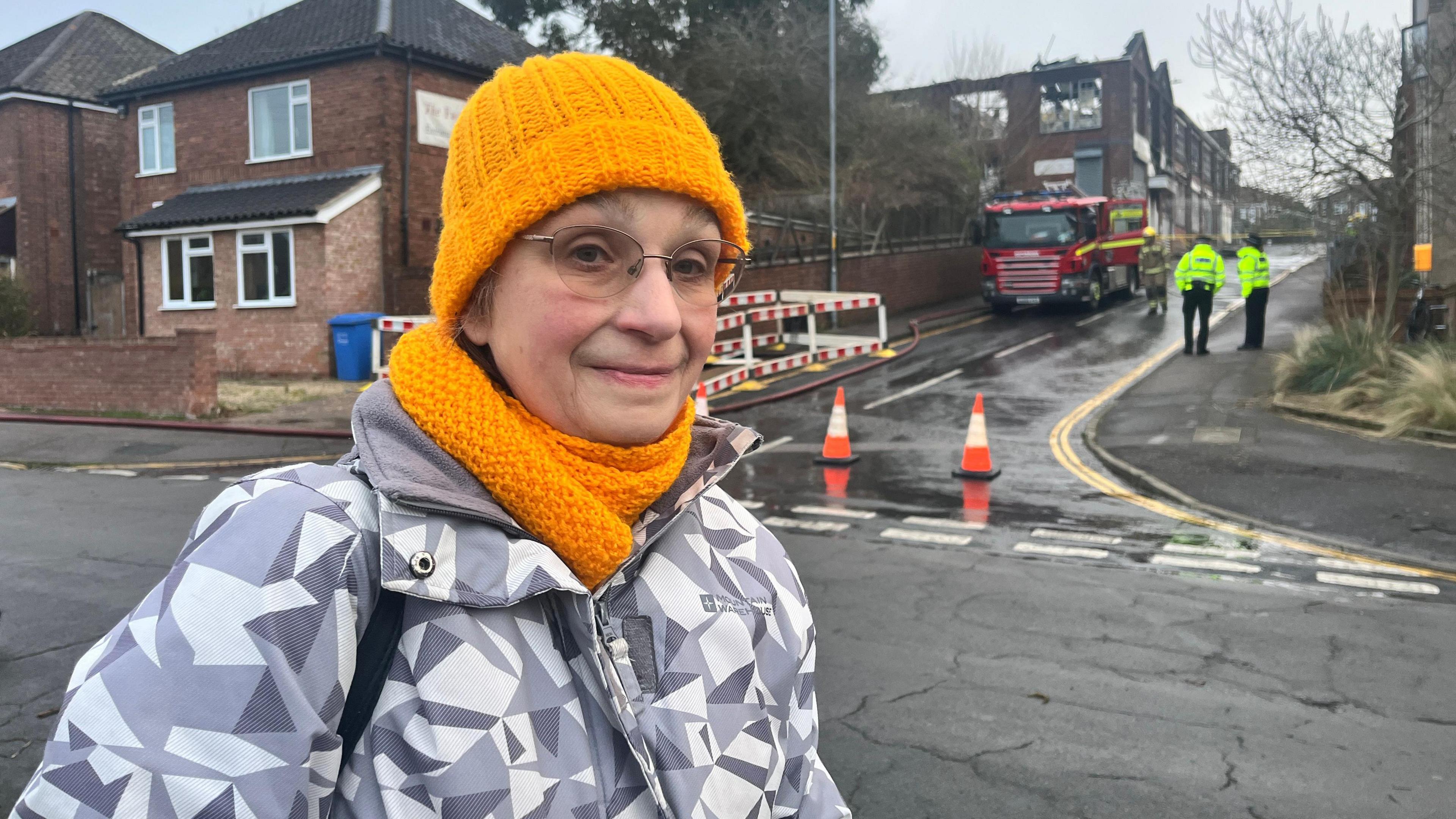 A woman wearing a matching orange knitted hat and scarf is smiling at the camera. There is a fire engine and emergency workers in the photo.