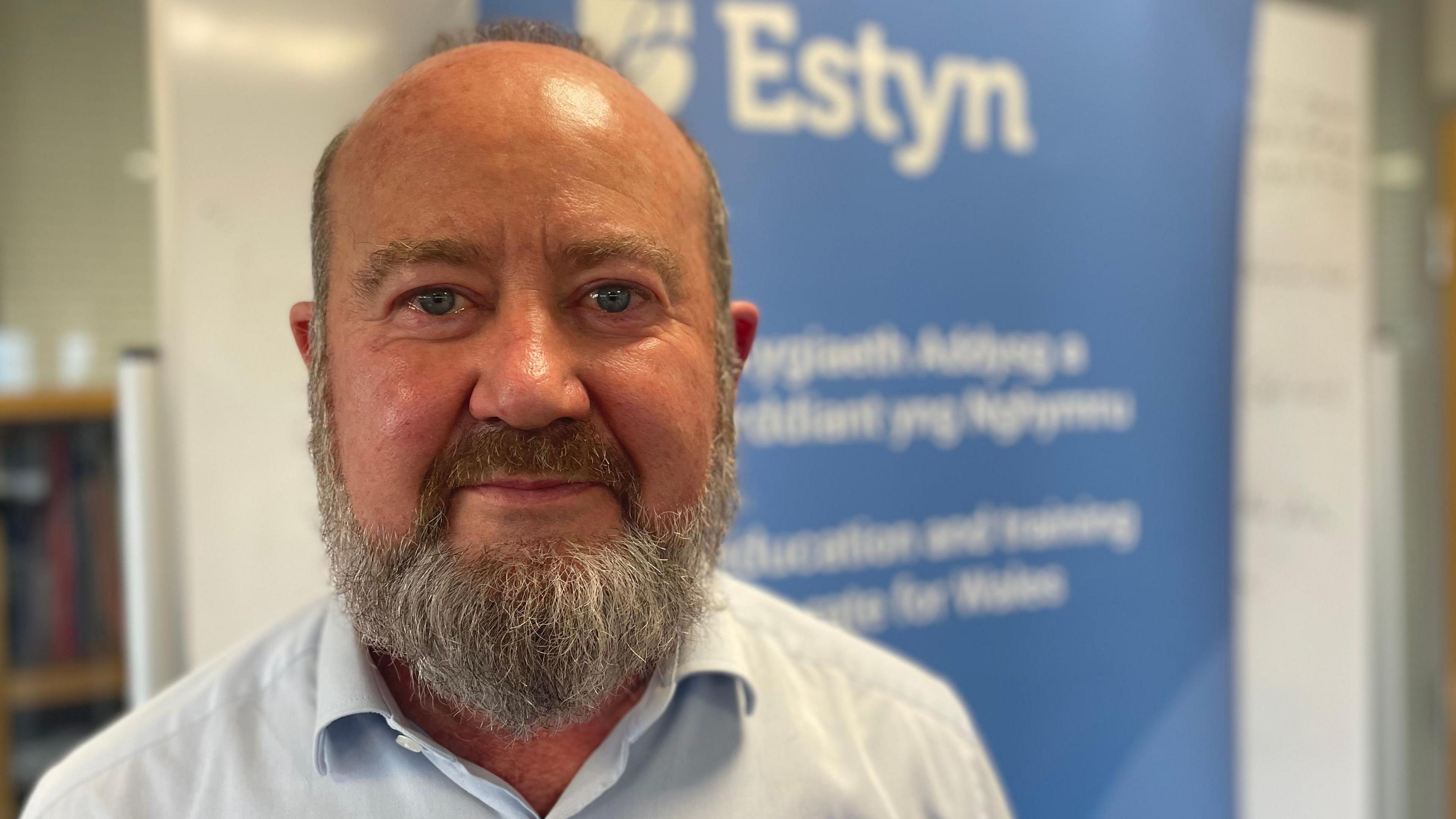 Owen Evans is looking into the camera and standing in front of an Estyn sign. He has a grey beard and is wearing a blue shirt. 