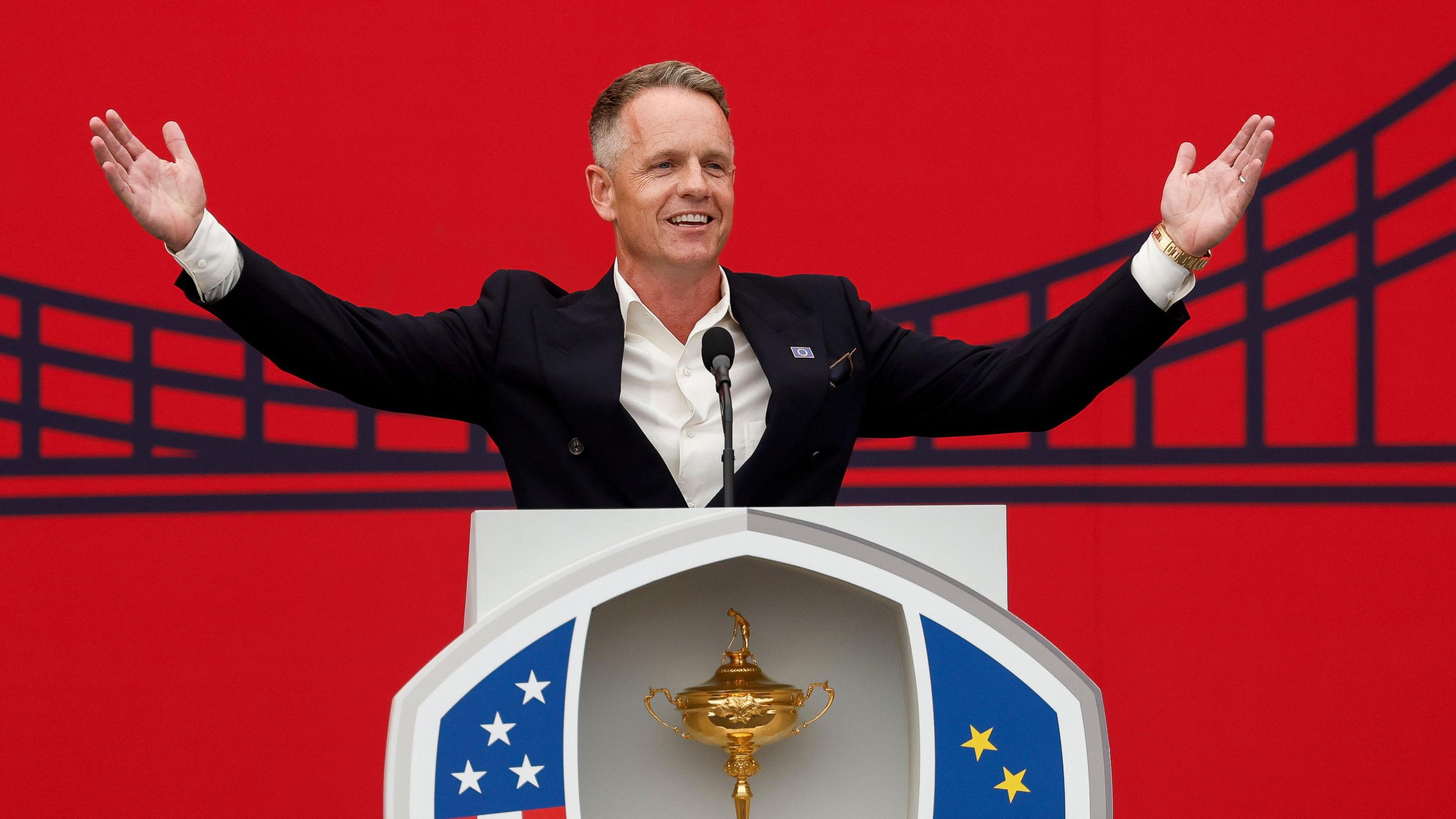 Europe captain Luke Donald stands with his arms outstretched behind the Ryder Cup and a microphone