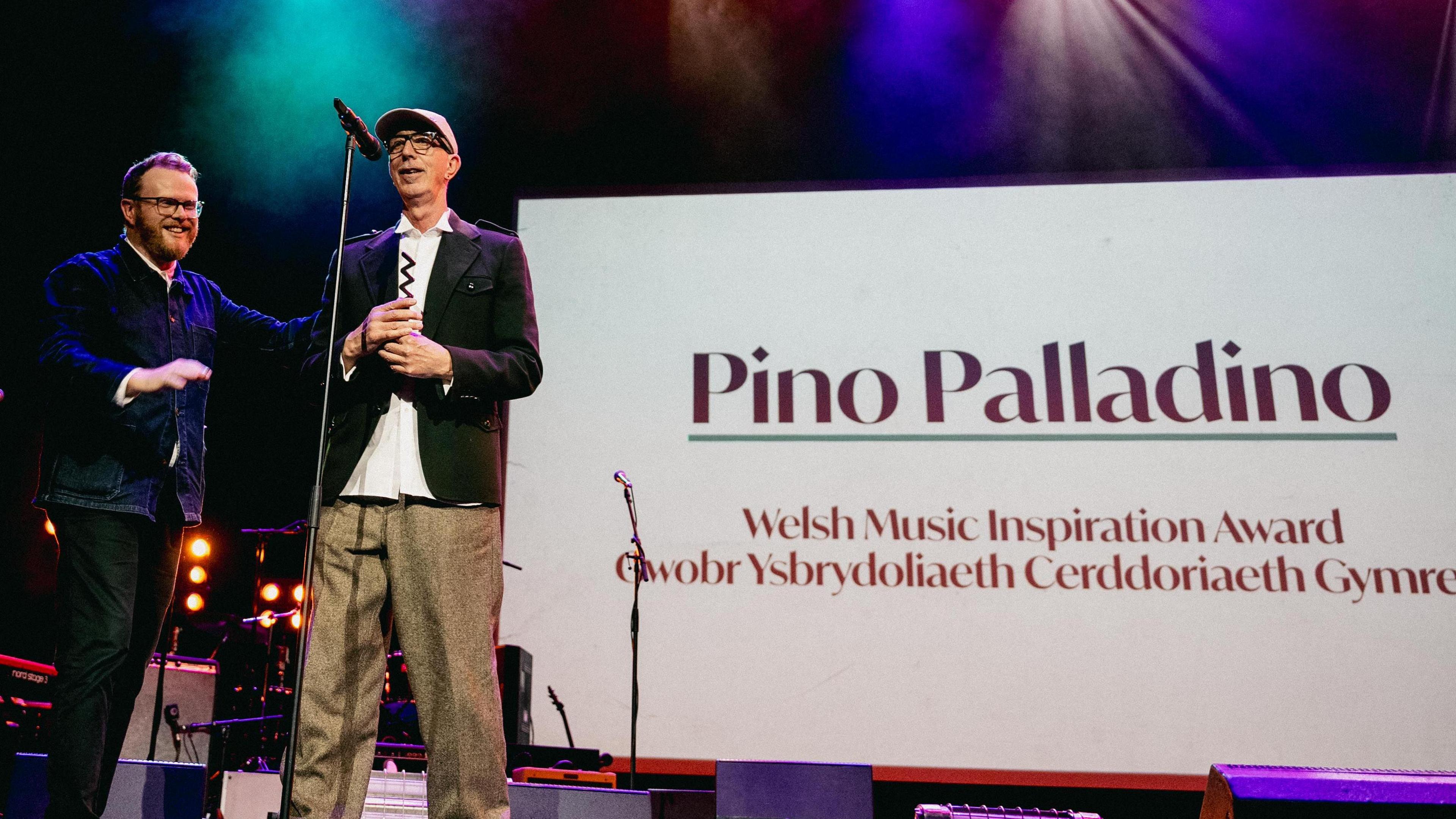 Pino Palladino wearing a white shirt, black jacket and brown trousers. He is standing on the stage with Huw Stephens. Behind him there is a screen that says 'Pino Palladino, Welsh Music Inspiration Award'. It is translated in Welsh too. 