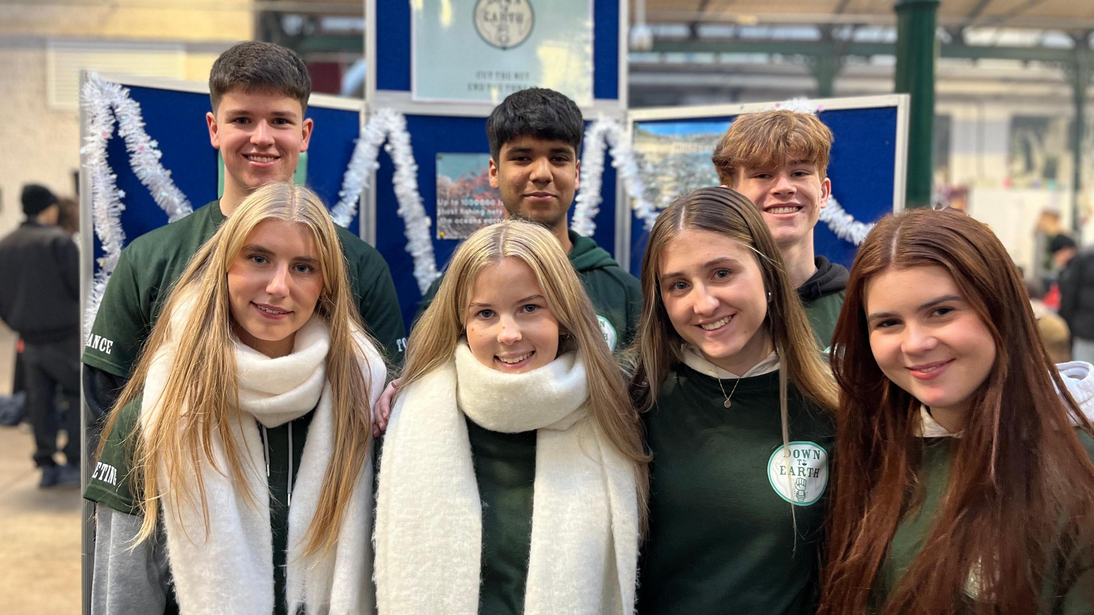 Seven members of the student company Down to Earth. Four girls at the front and three boys at the back all smiling at camera. They are wearing green t-shirts with the brand name printed on the front. Two girls are wearing white scarves. Stall board is in the background. 