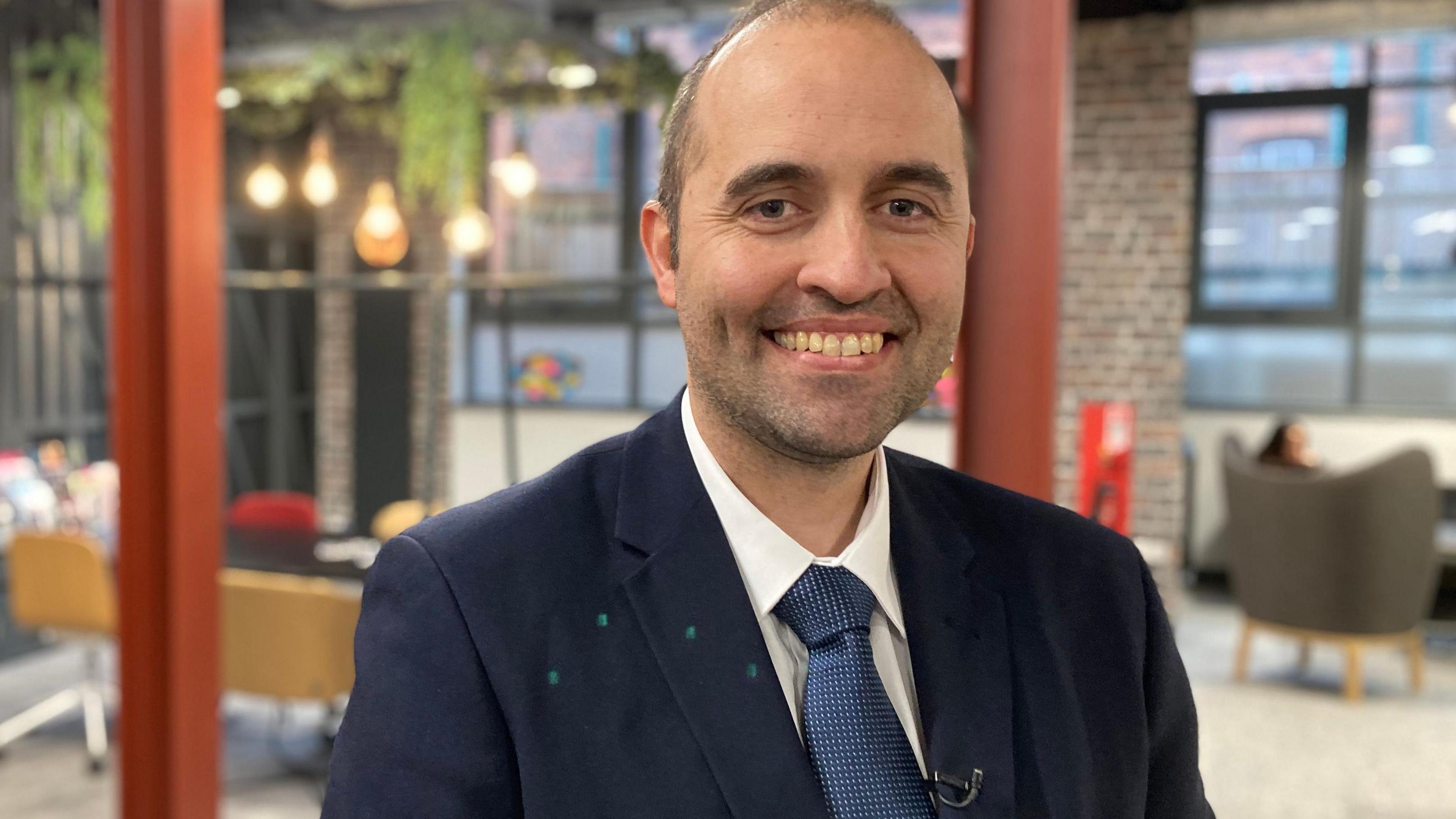 A man wearing a blue jacket with blue tie and white shirt smiling to the camera. He has short brown hair and it is receding.