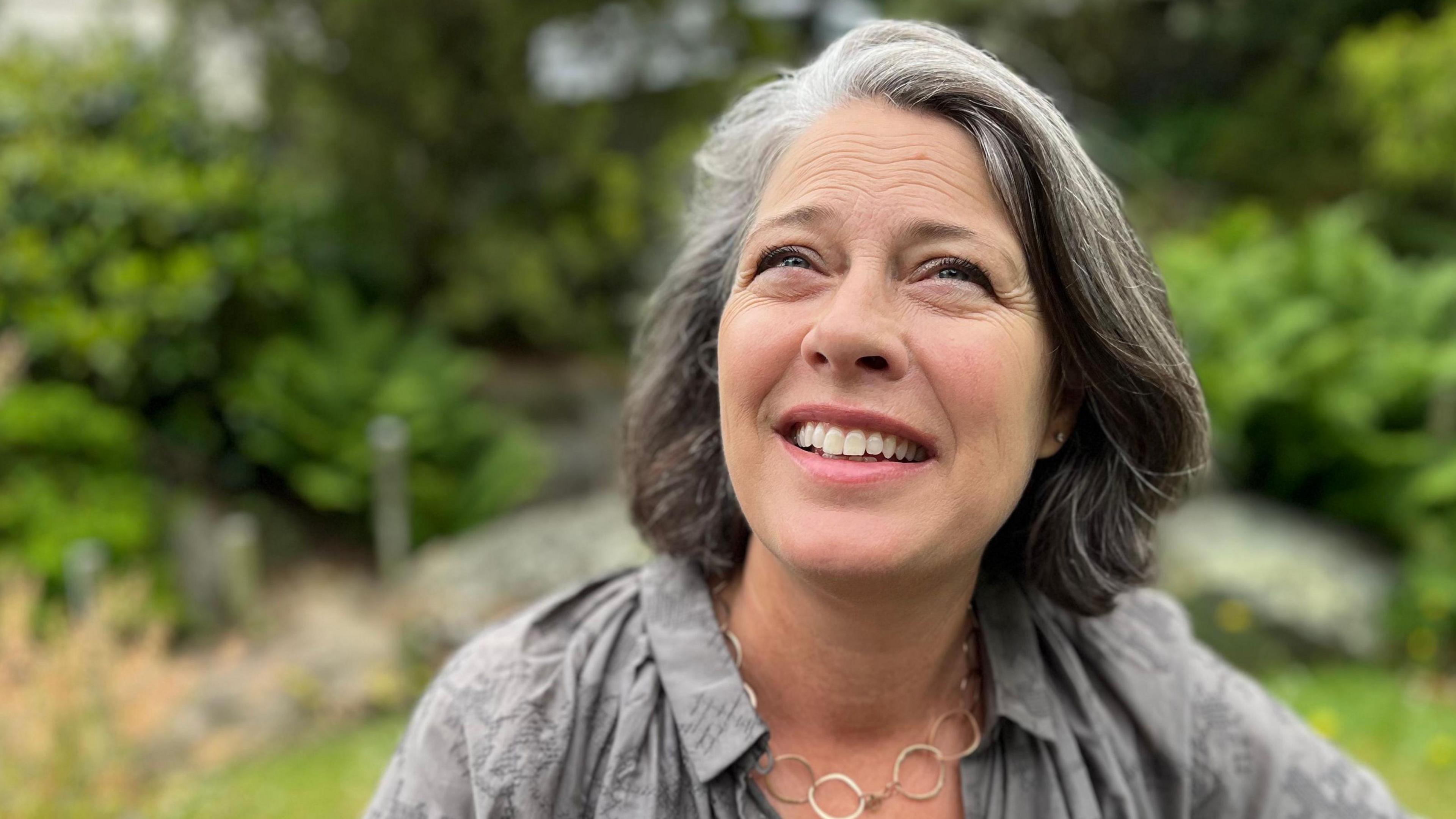 A lady with shoulder length grey hair looking towards the camera. She's wearing a grey blouse and a necklace with metal hoops. 