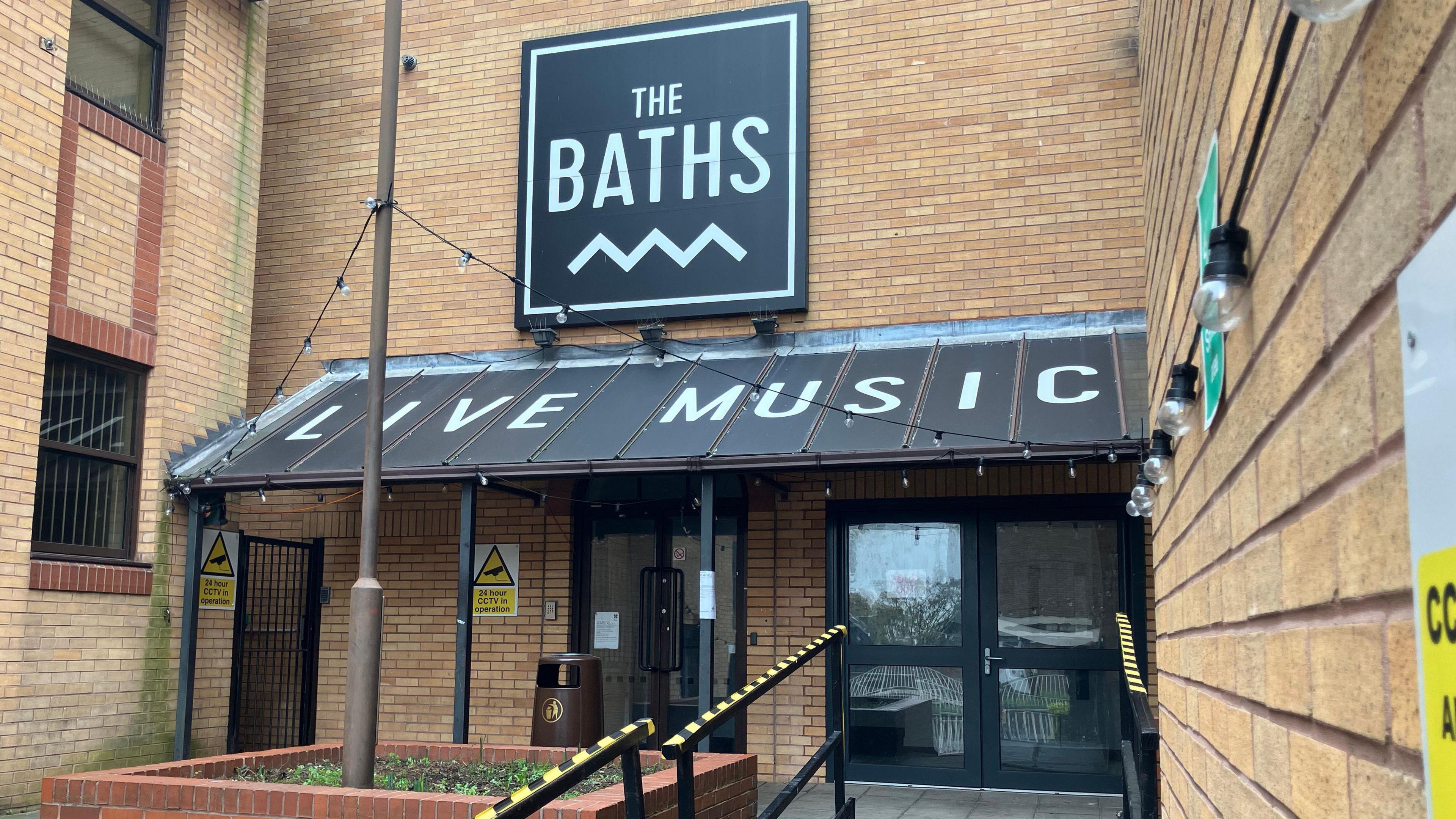 A general view of the entrance to The Baths in Ipswich. A black sign above the entrance reads 'The Baths' in white writing with a cover over the doors that reads 'Live Music' in white writing.
