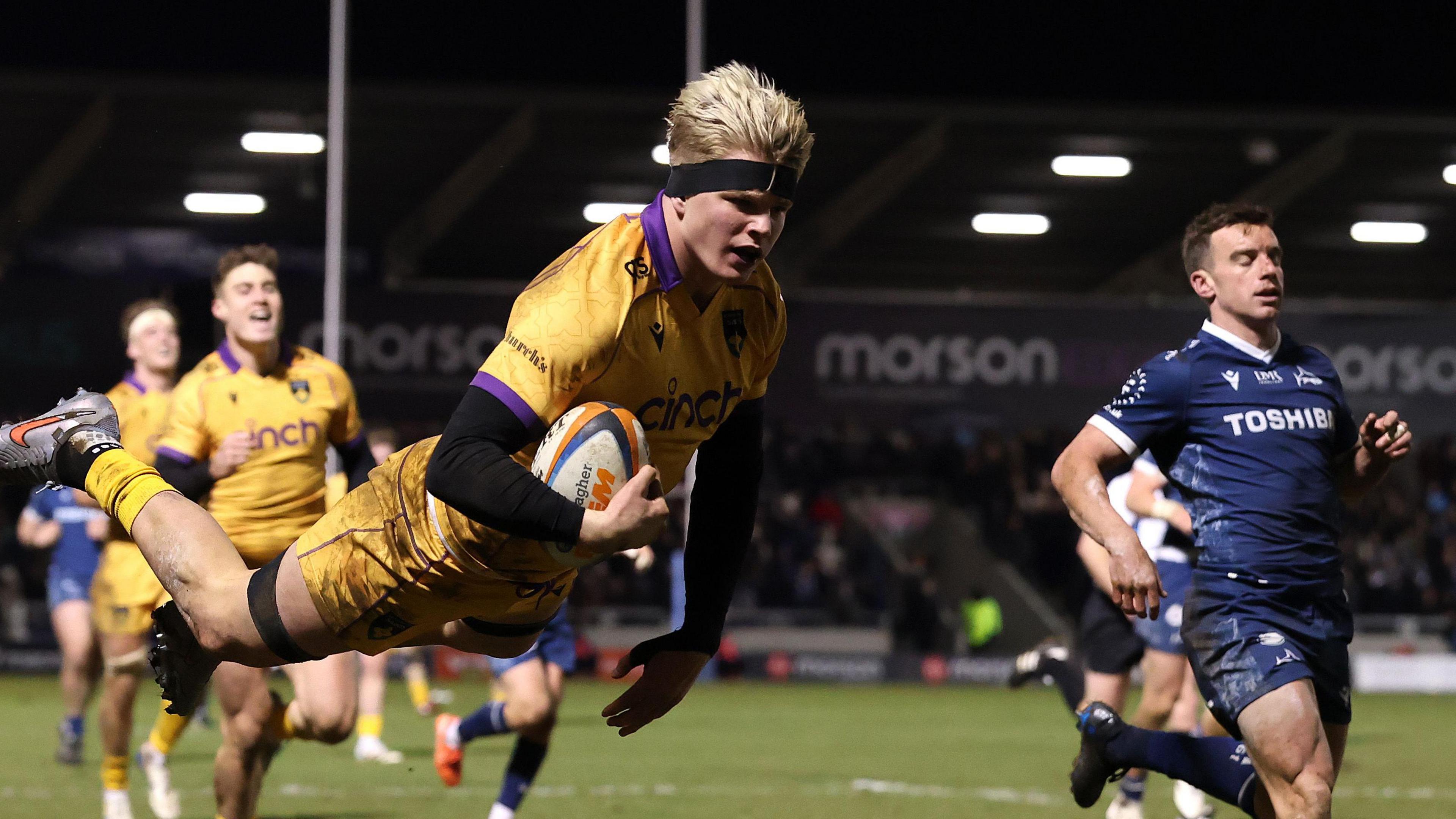 Henry Pollock, wearing a black headband, leaps in the air with the ball under his arm, to score Northampton's final try as Sale fly-half George Ford watches on