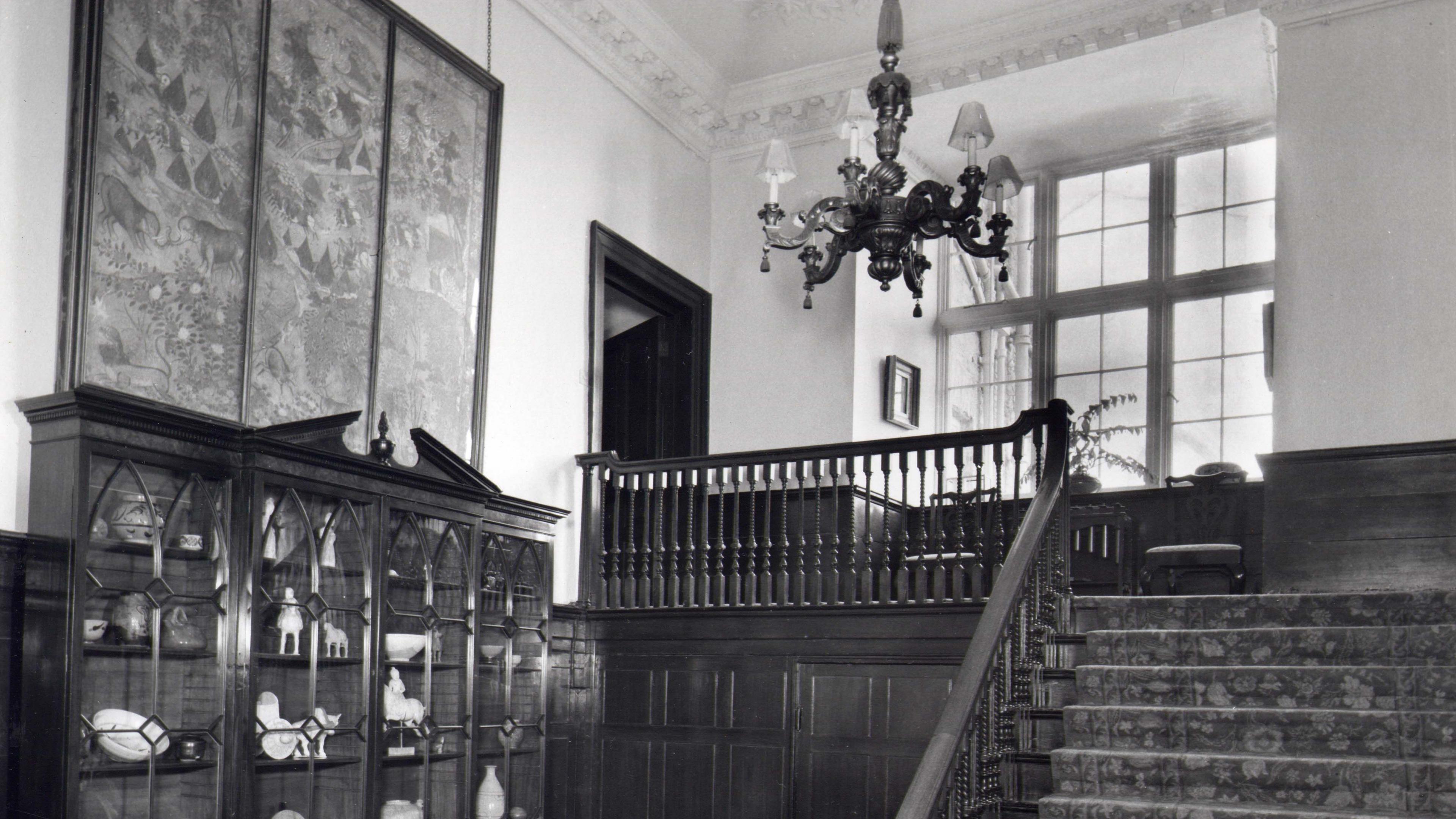 A black and white photo shows an ornate handrail and stairs that lead down into a hallway with a chequered, tiled floor. There is a dark wooden dresser again a wall, displaying china.