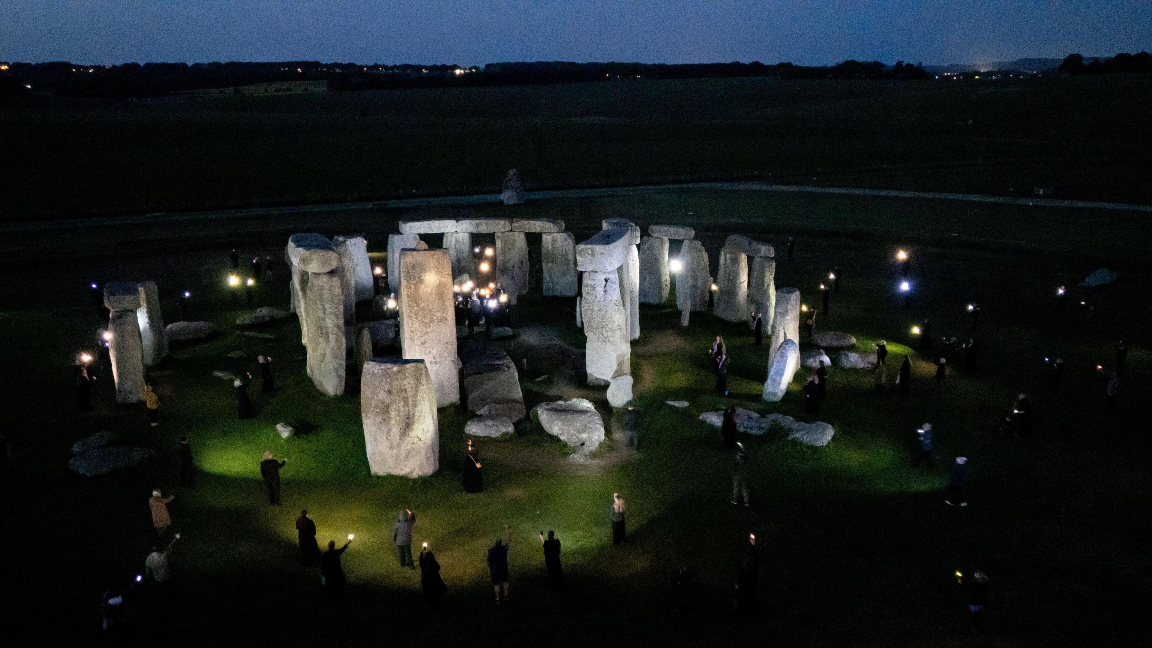 A drone shot of Stonehenge at night showing people standing round it in the dark with lanterns. The stones are lit up by floodlights.