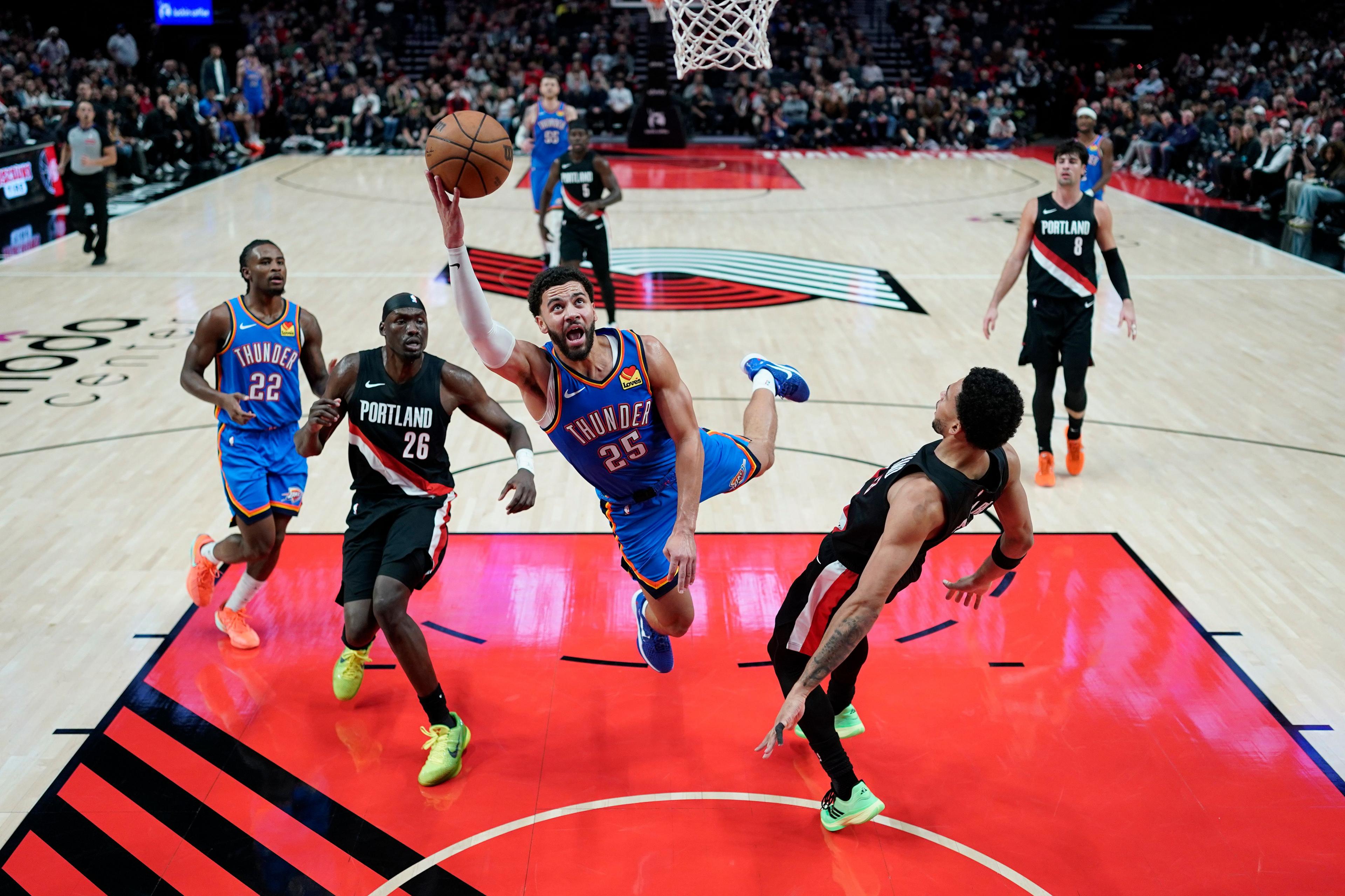 Basketball player in a blue Thunder jersey leaps toward the hoop for a layup while holding the ball, surrounded by Portland defenders in black uniforms on a brightly lit court with the Trail Blazers logo visible and spectators in the background.