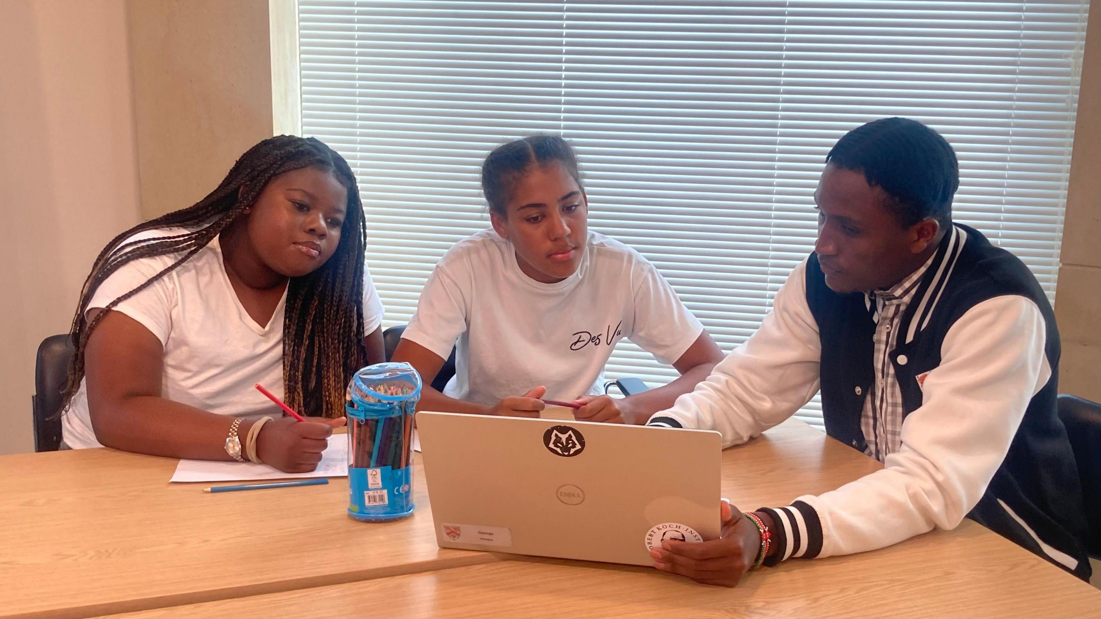 Two girls in white T-shirts are leaning on a table in a room, holding pencils or pens, looking at a laptop screen, with George Wanjiru to their left.