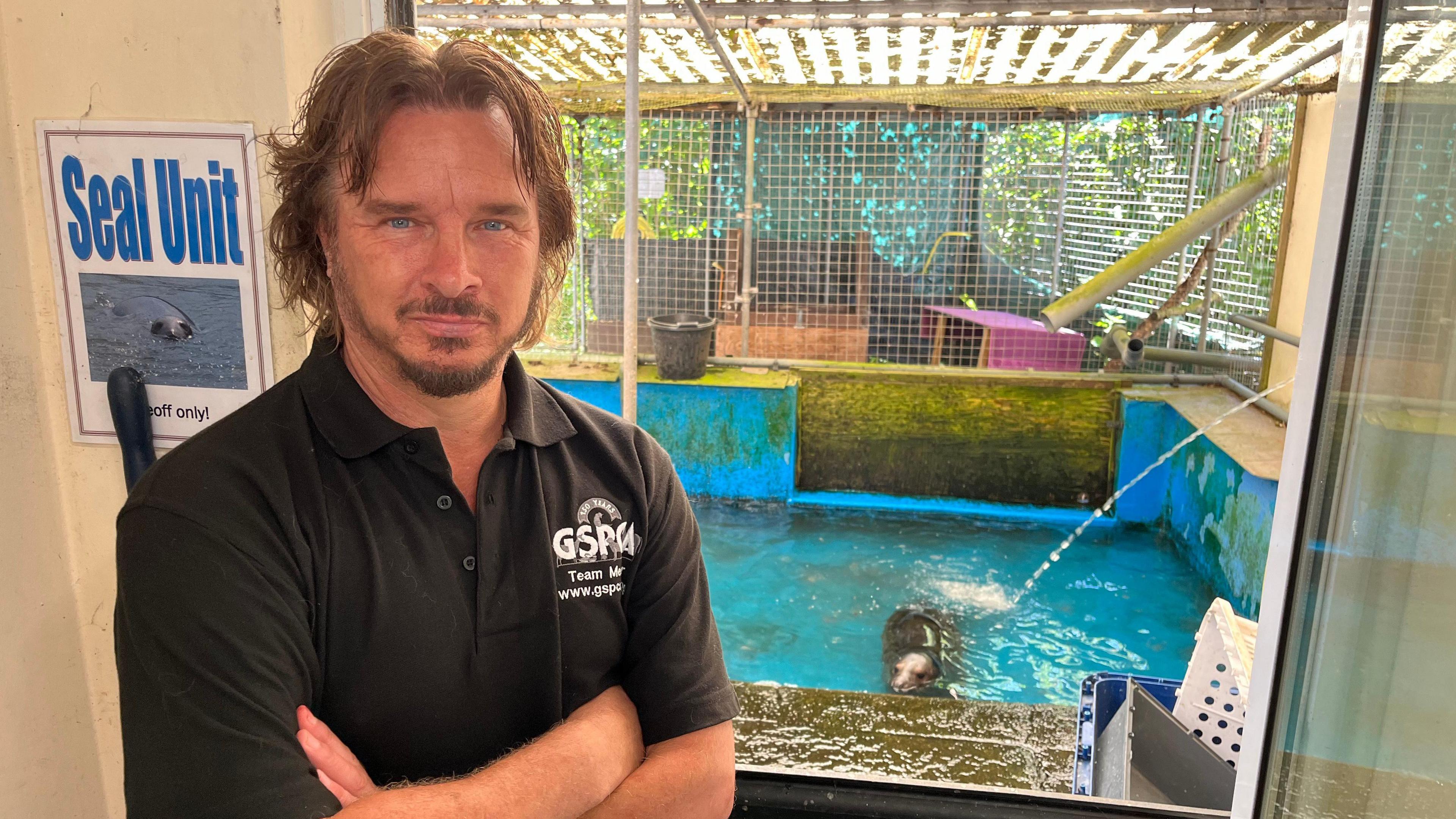 A man in a t-shirt that says GSPCA  standing in front of a swimming pool with a seal in it.