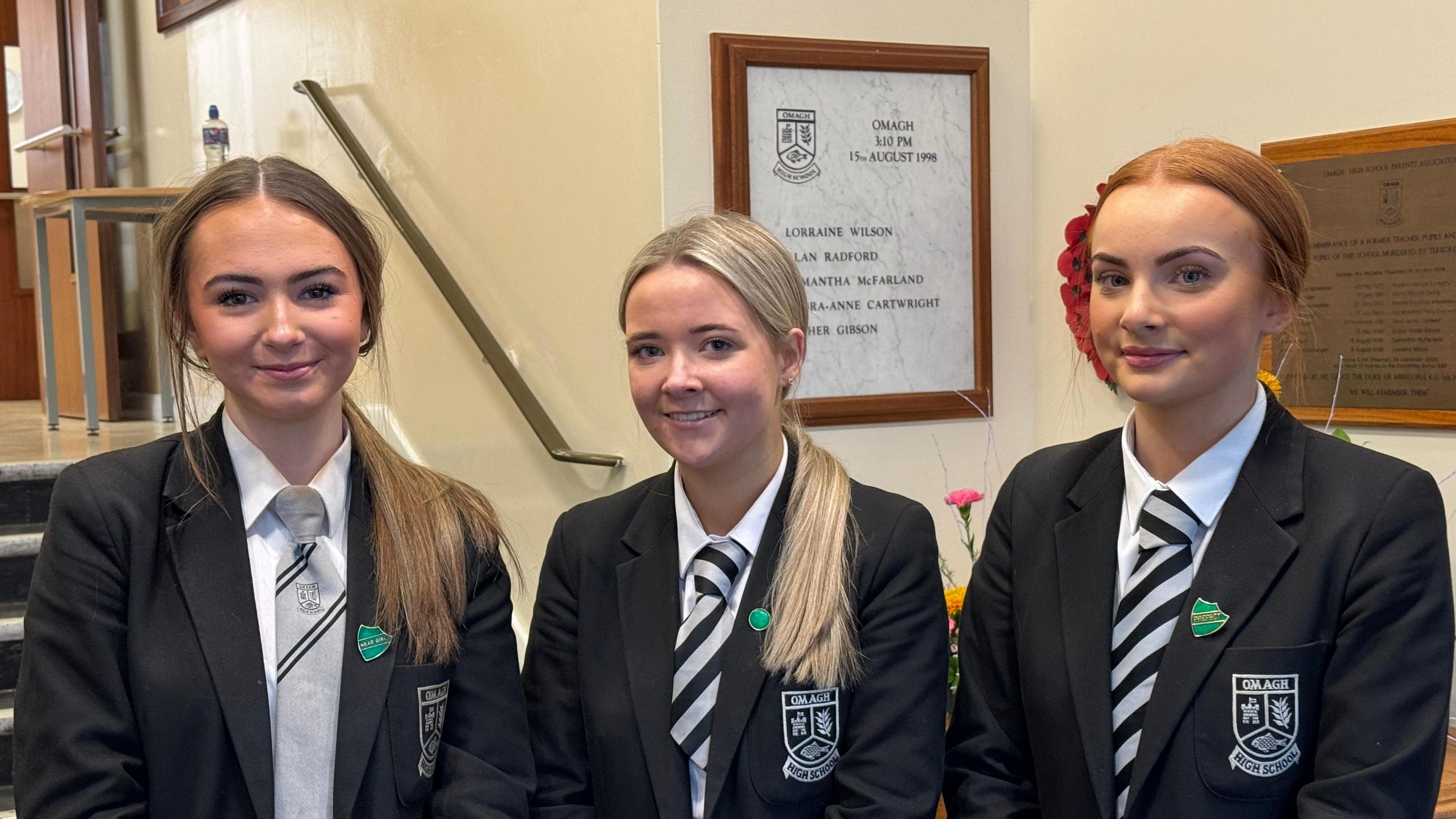 Omagh High School pupils Poppy, Connie, and Louise. All have ponytails, Poppy has brown hair, Connie has blonde hair, and Louise has red hair. They are all wearing their school uniforms - black blazer, white shirt and black and grey tie.