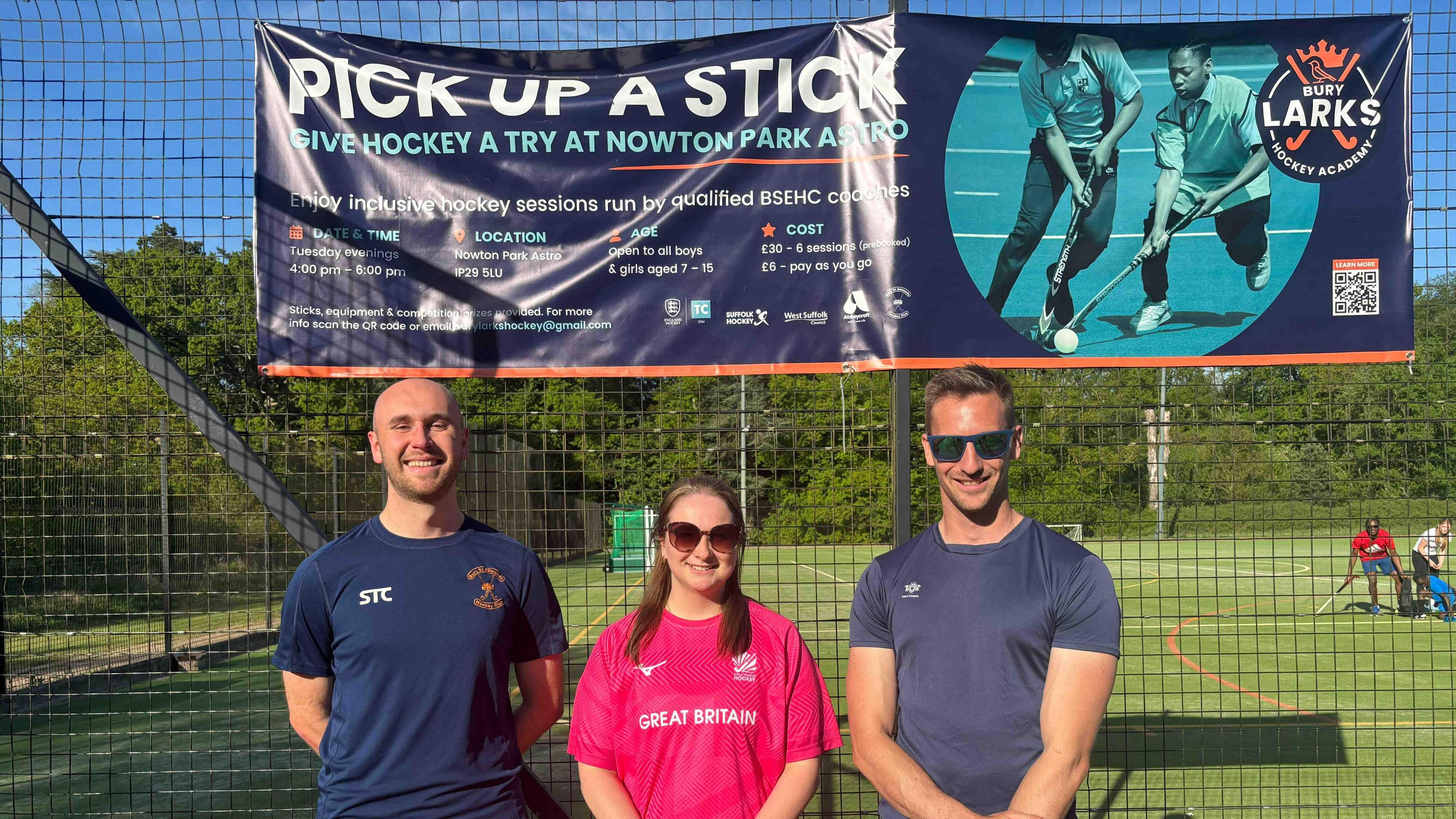 Three hockey coaches stand in front of the pitch. Finn and Harry stand either side of Emily, dressed in navy blue sports attire while Emily is wearing a pink hockey top.