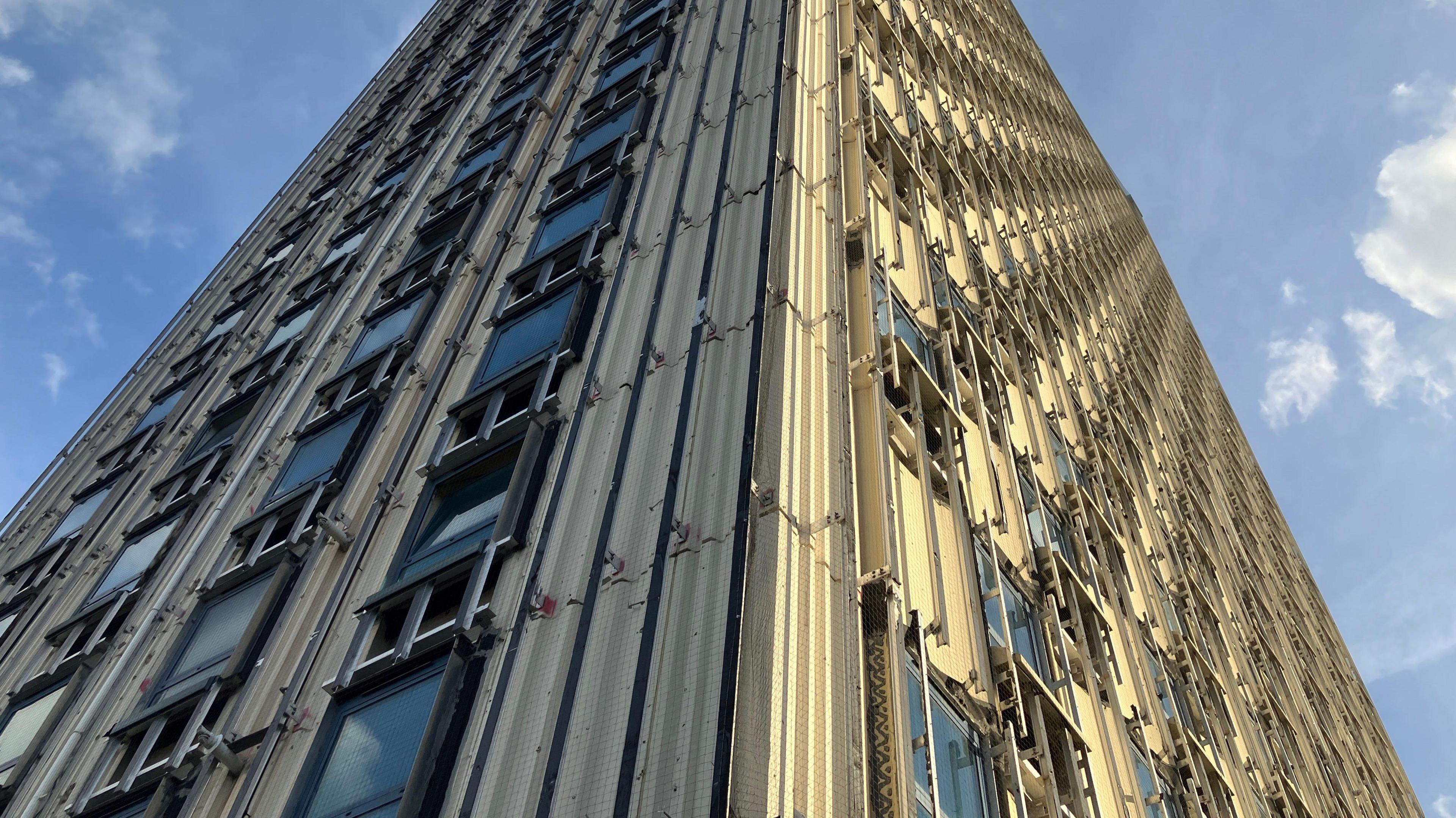 A close-up shot of Walbrook House, taken from below, showing the floors rising into the sky and rows of windows. It appears some of the concrete panels have been removed from the building's facade