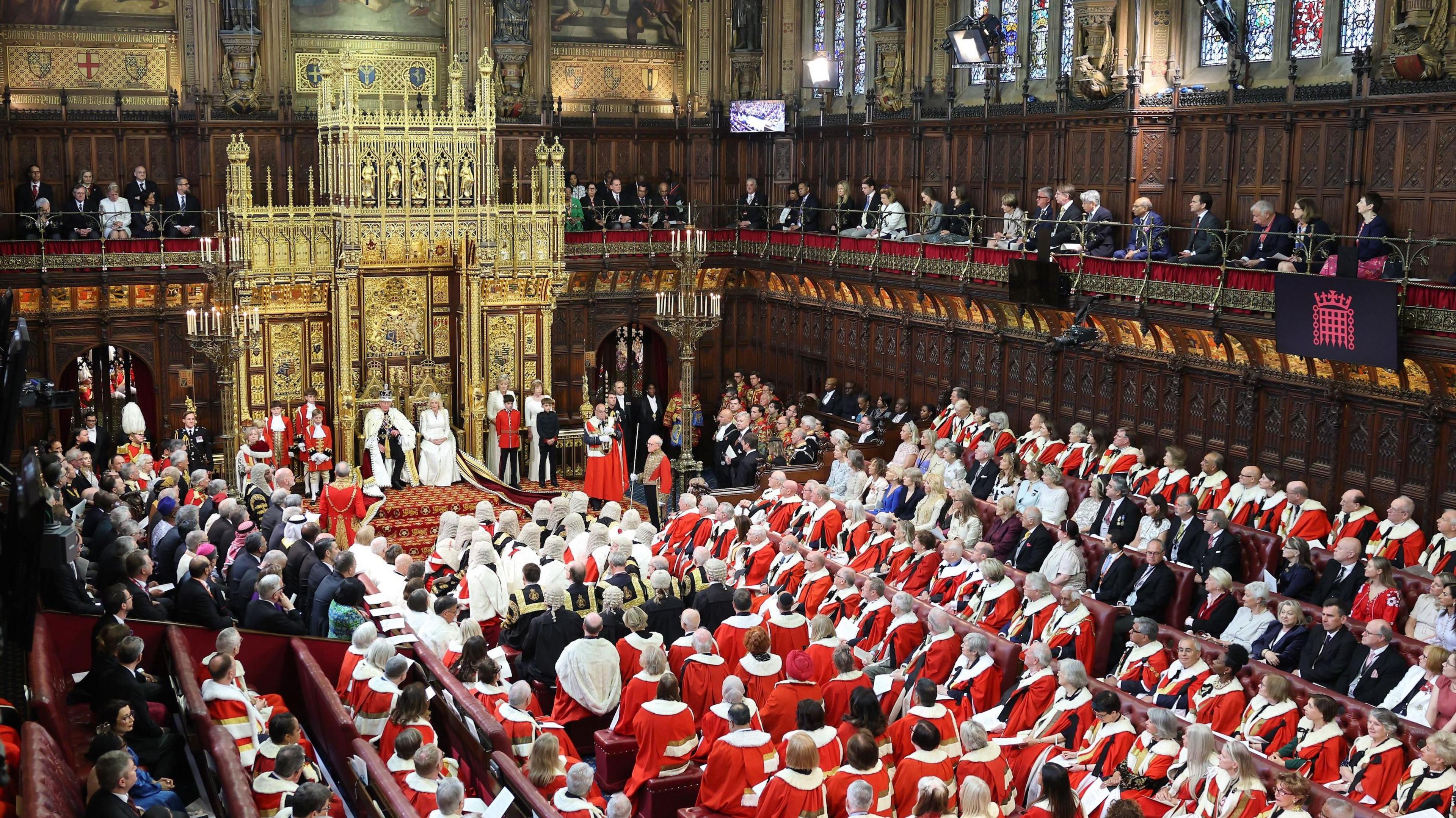 Birds Eye view of the kings speech, at the far end of the room you can see King Charles and queen camilla in fancy robes and crowns, and there are lots of people wearing red and white outfits sitting looking at them