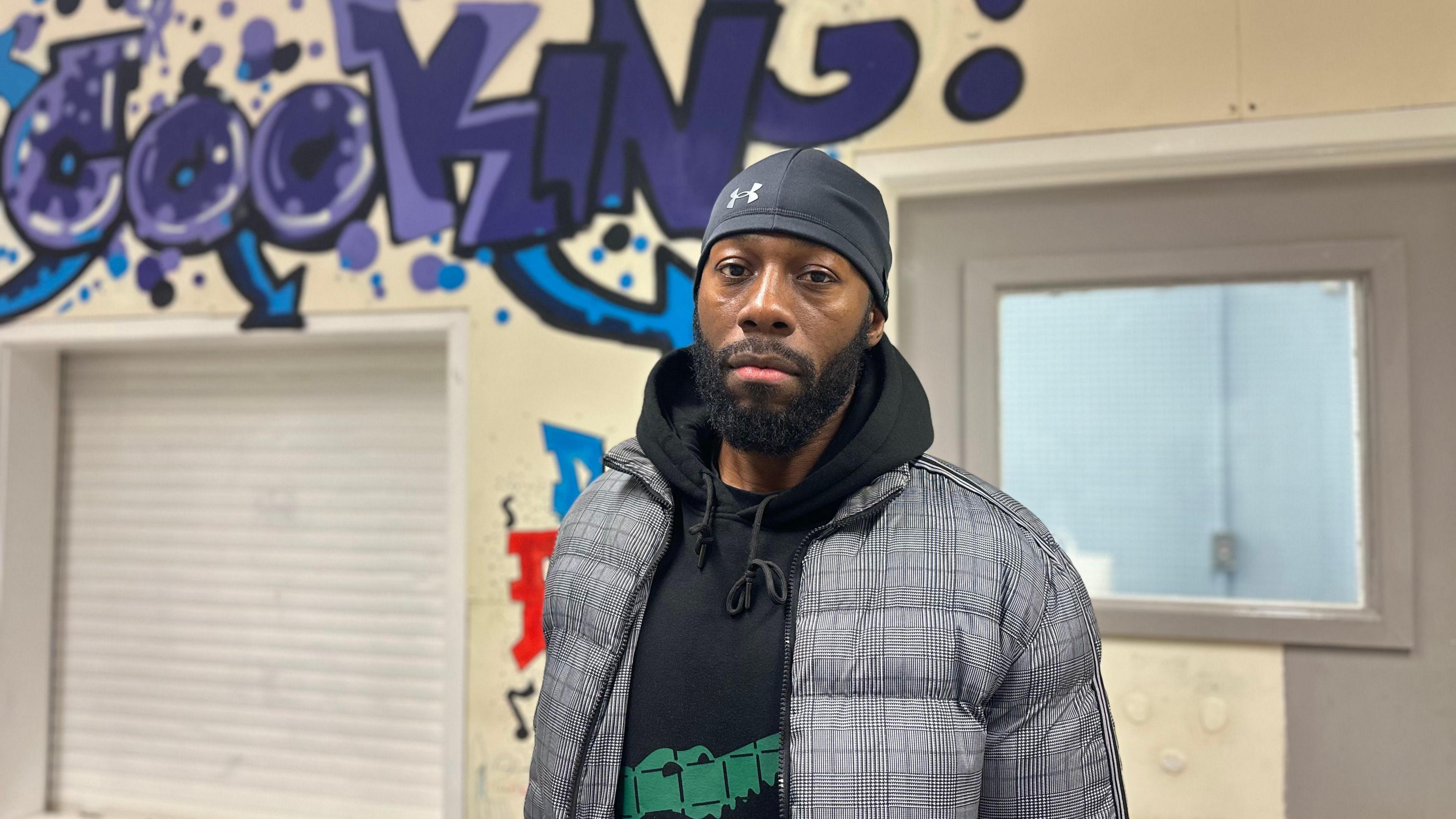 A man wearing a grey durag, a black hoodie and a grey patterned coat stands in a cream-coloured room with purple graffiti in the background that reads "cooking".
