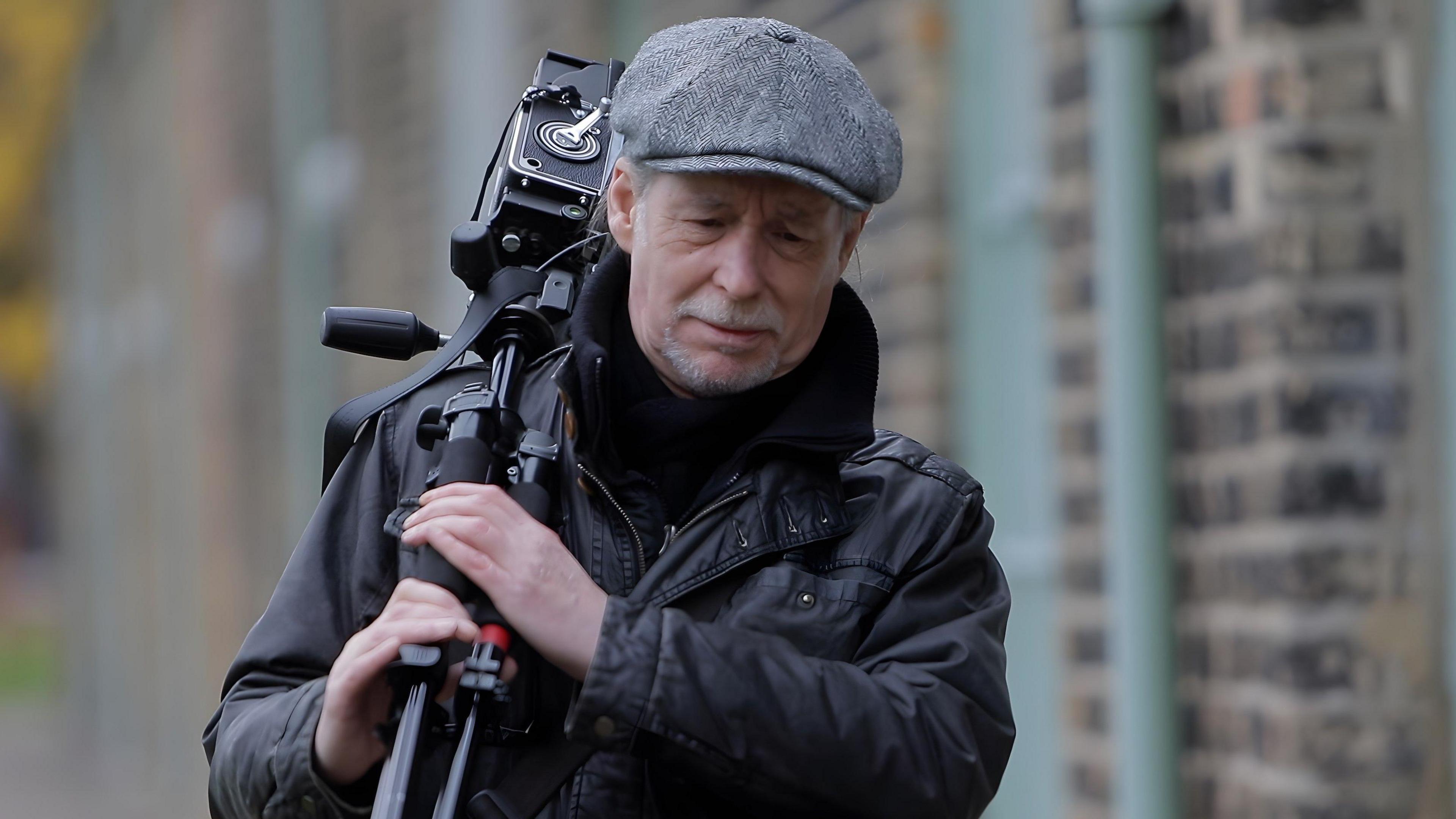 Mik Critchlow is carrying a camera over his shoulder. He has grey stubble and is wearing a black coat and has a grey cap on. He is walking alongside a row of cottages 