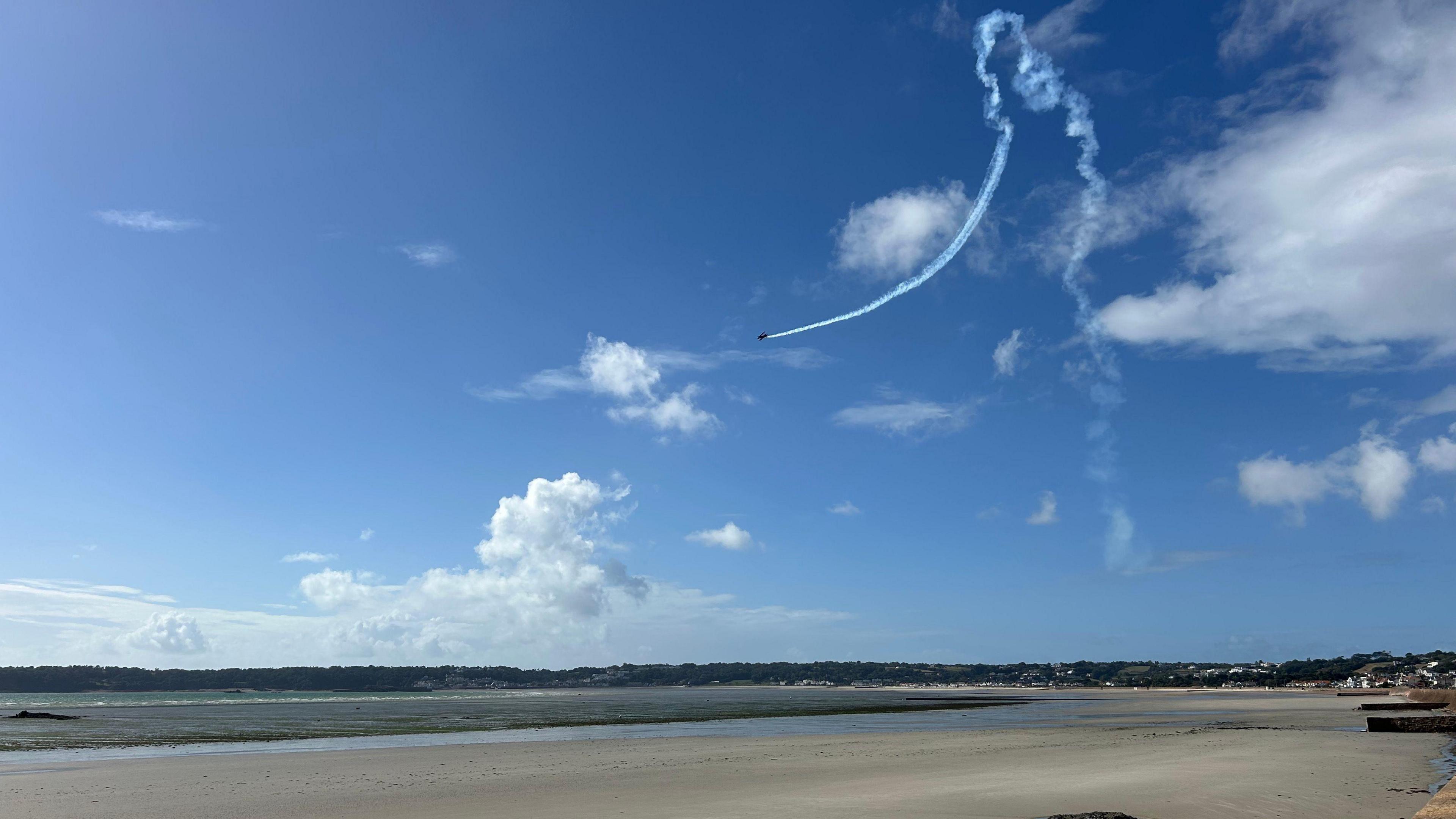 A plane in the sky above a wide sandy beach. A smoke trail behind shows it has done a sharp manoeuvre.