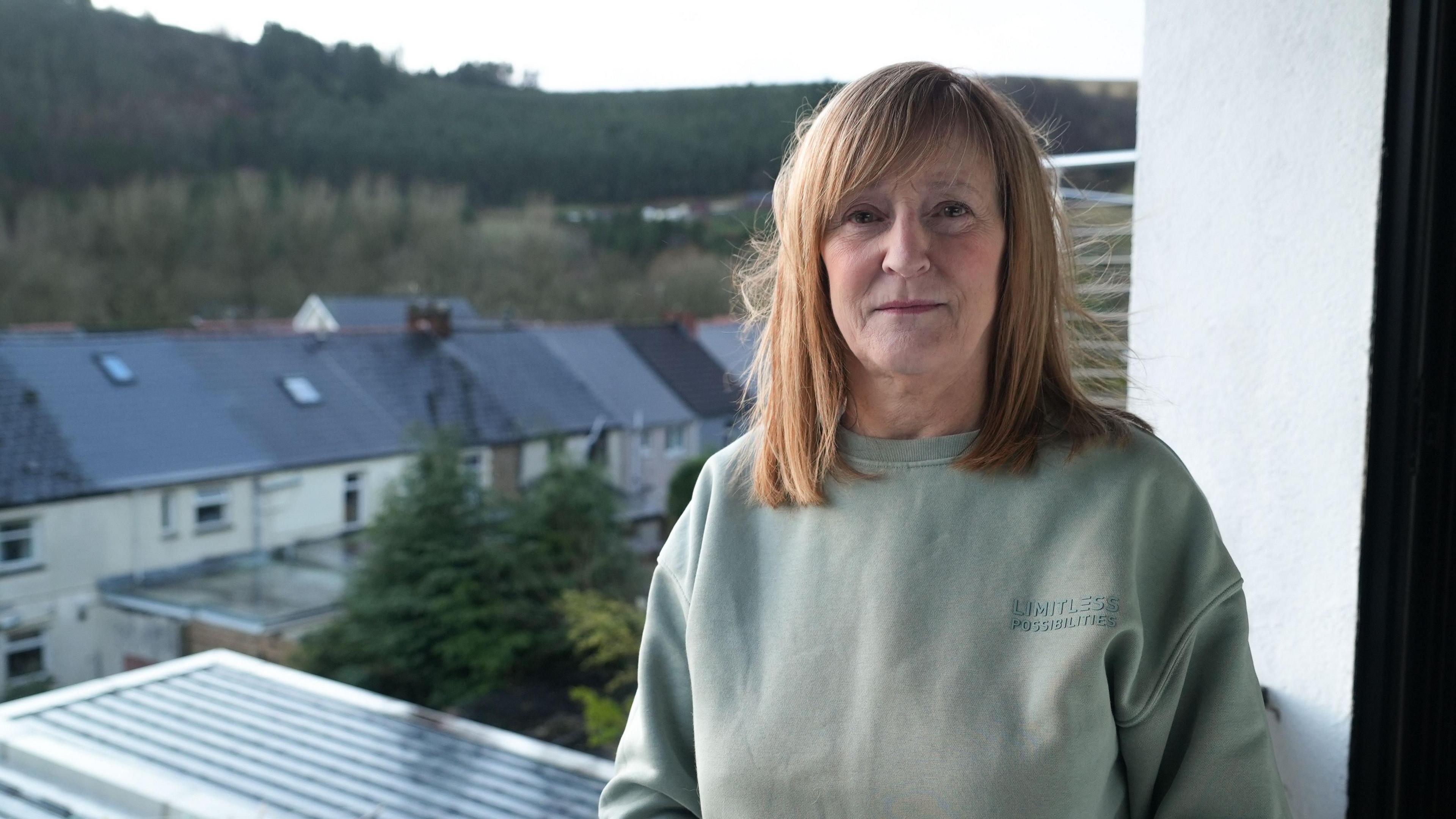 Dianne standing in a window of her home, which looks out onto trees. she is wearing a green cotton jumper and has red shoulder length hair.