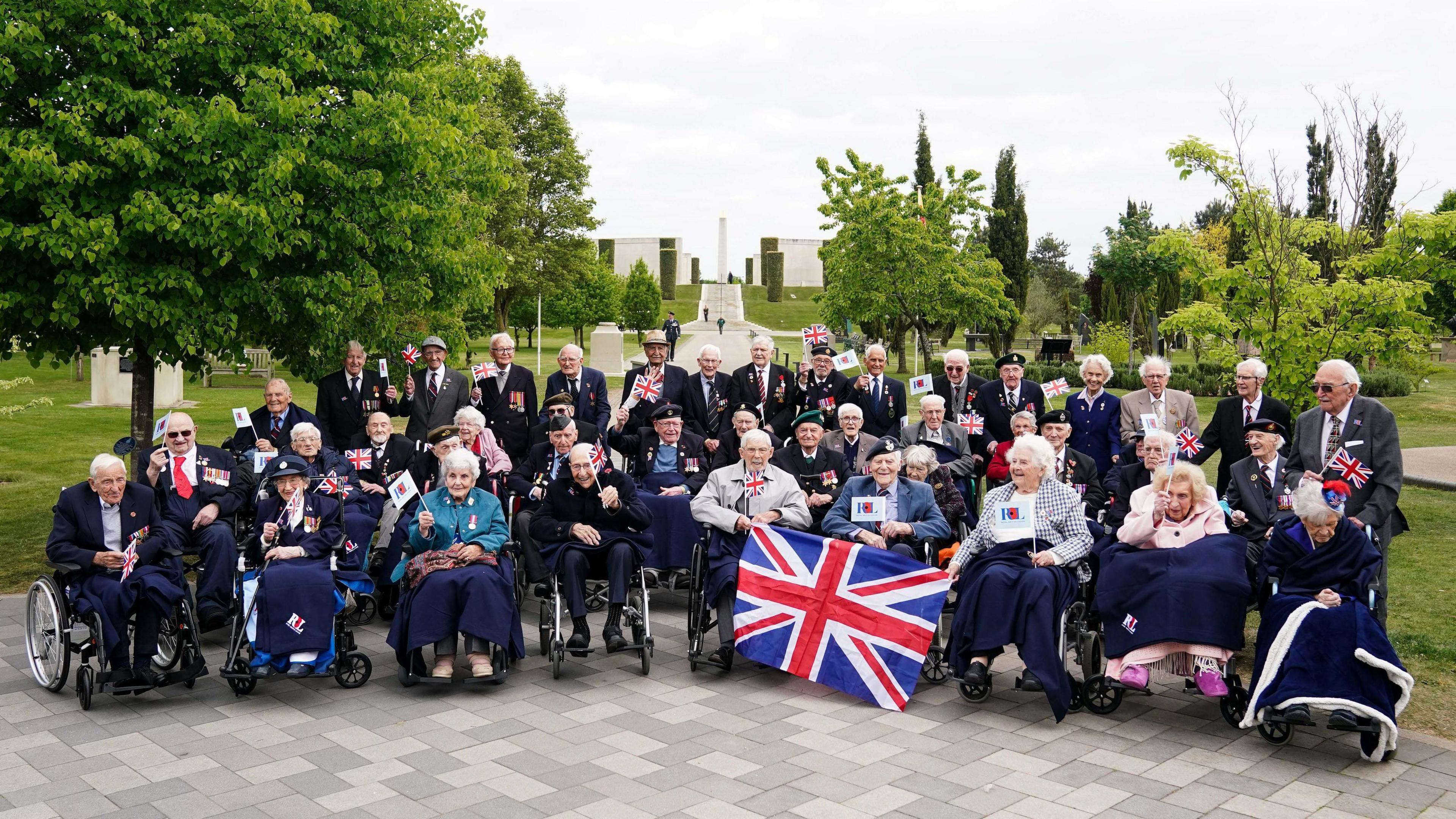 A number of veterans, some sitting in wheelchairs and holding union jacks, are gathered on a pavement with a memorial visible in the background.